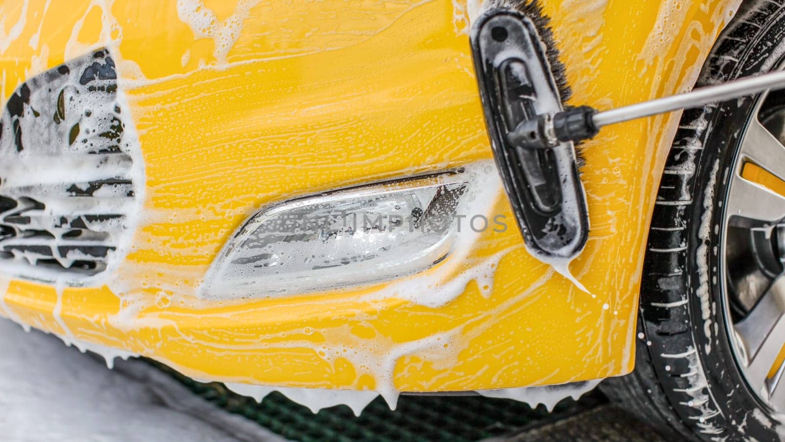 Brush cleaning front bumper and light covered with shampoo and foam, of car in carwash, 
