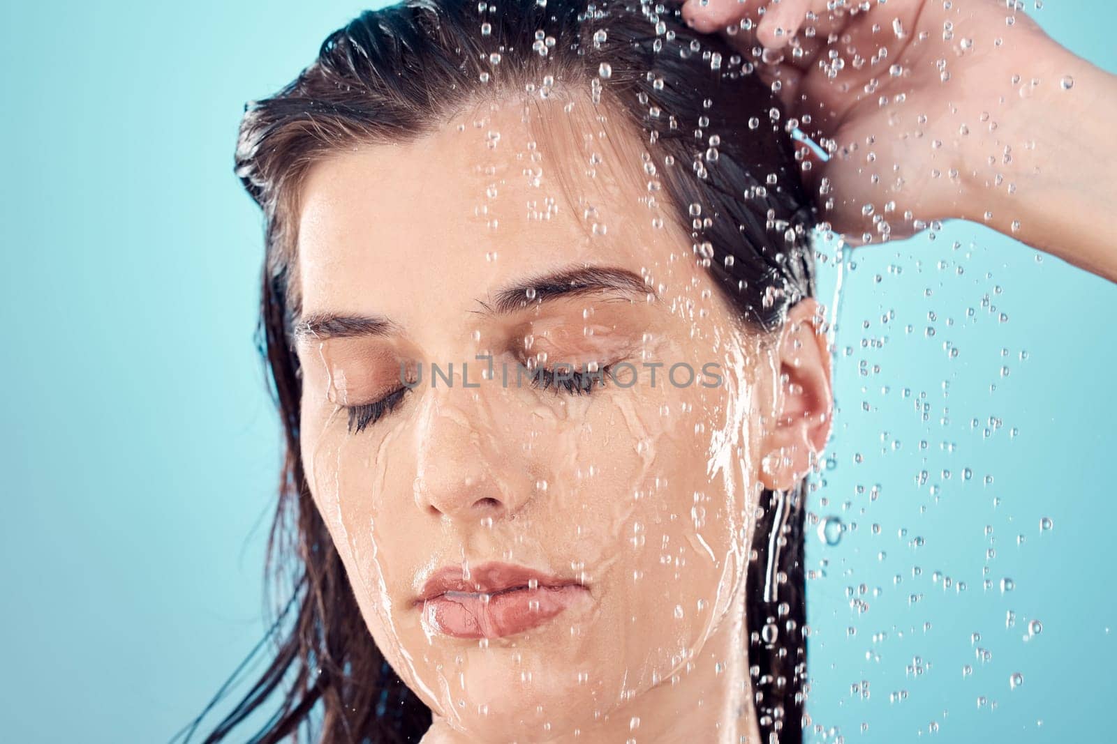 Water splash, hair care and face of woman in shower in studio isolated on a blue background. Beauty, eyes closed and young female model washing, cleaning or bathing for hygiene, skincare and health