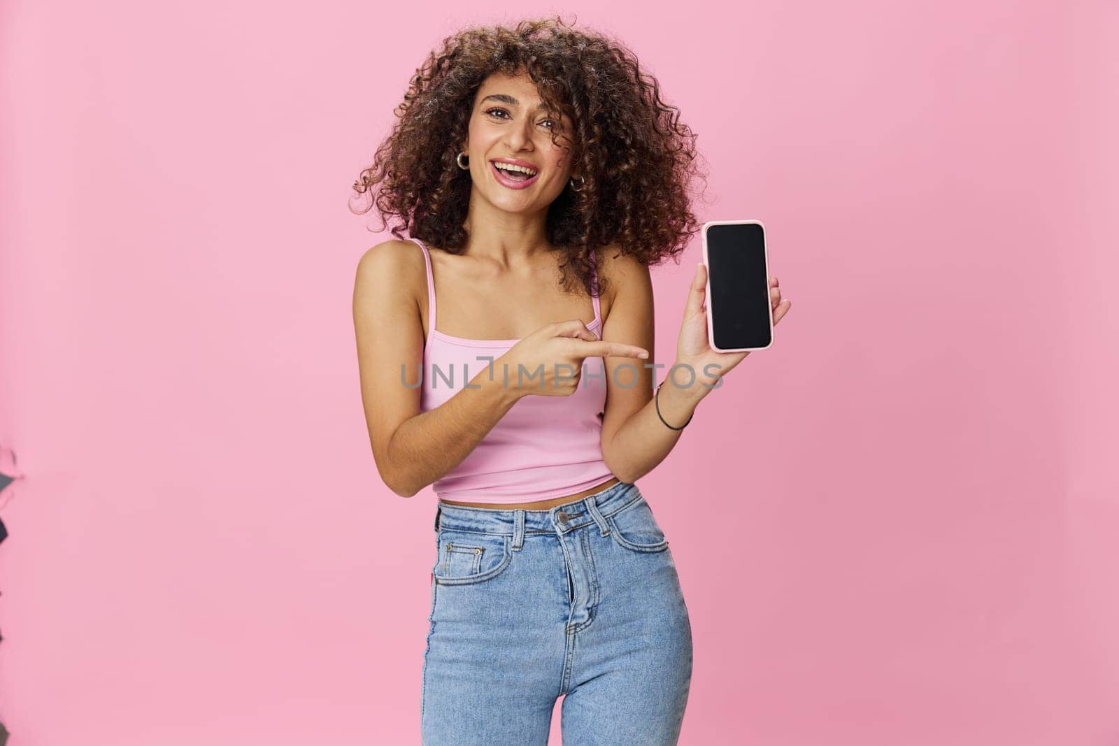 Woman blogger holding phone video call takes selfies, with curly hair in pink smile t-shirt and jeans poses on pink background, copy space, technology and social media, online. High quality photo