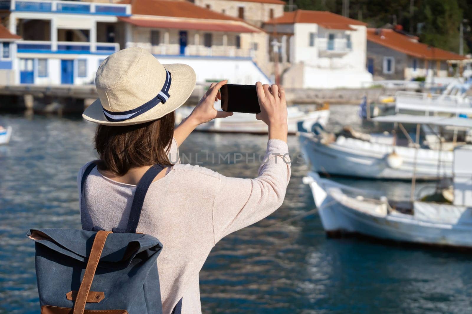A young girl in a hat and with a backpack makes a photo of a beautiful bay with yachts and boats on the sea while traveling, a tourist walks along the shore next to the ocean and takes pictures.