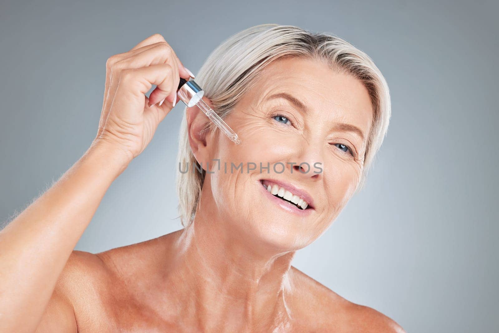 Senior woman, skincare and serum for beauty, cosmetics or anti aging treatment against a grey studio background. Happy elderly female applying oil with dropper and smile in satisfaction for skin.