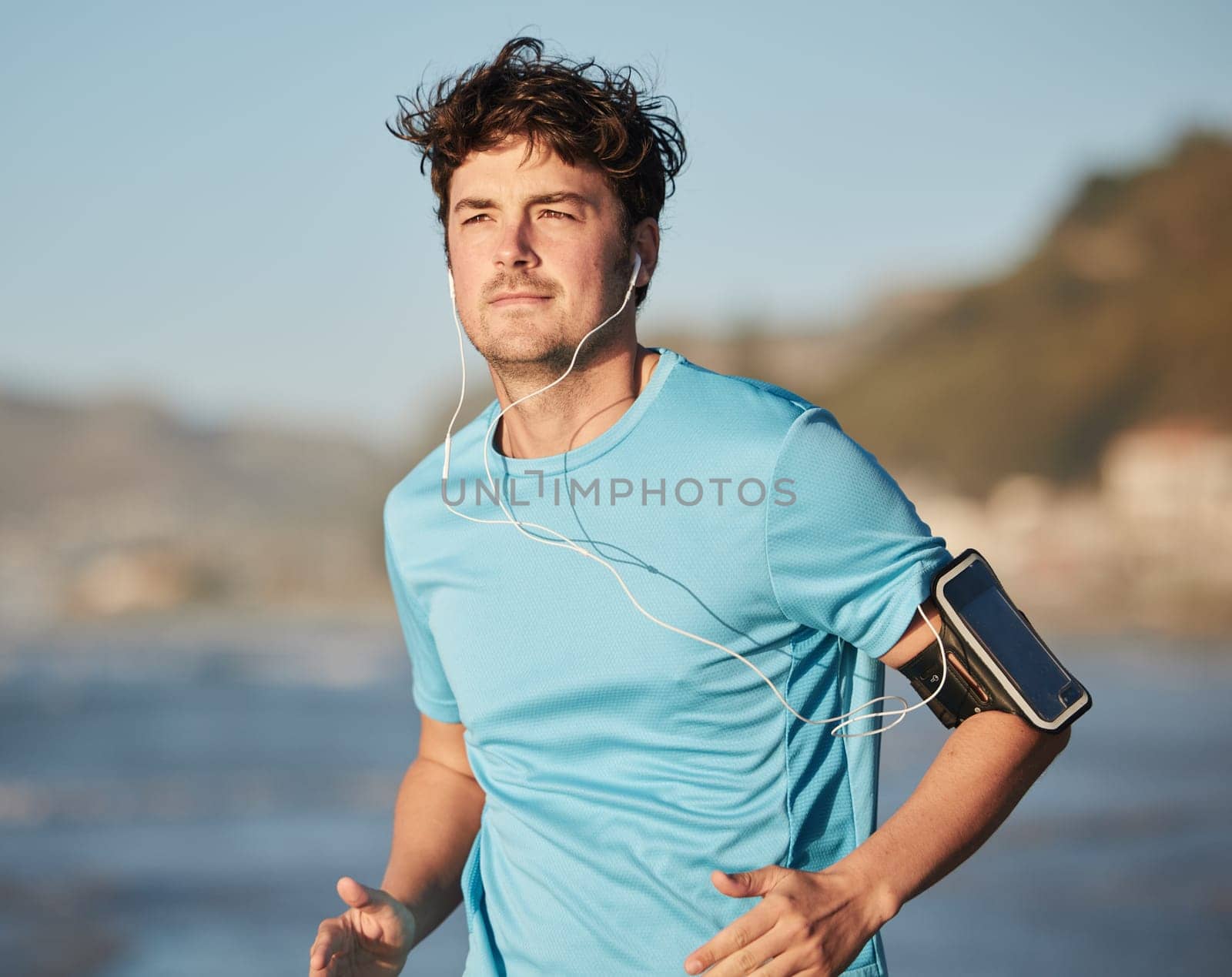 Running, music and man athlete at beach with headphones run by the ocean with audio streaming. Mobile radio, web podcast or training song of a runner athlete listening to a track on an outdoor run.