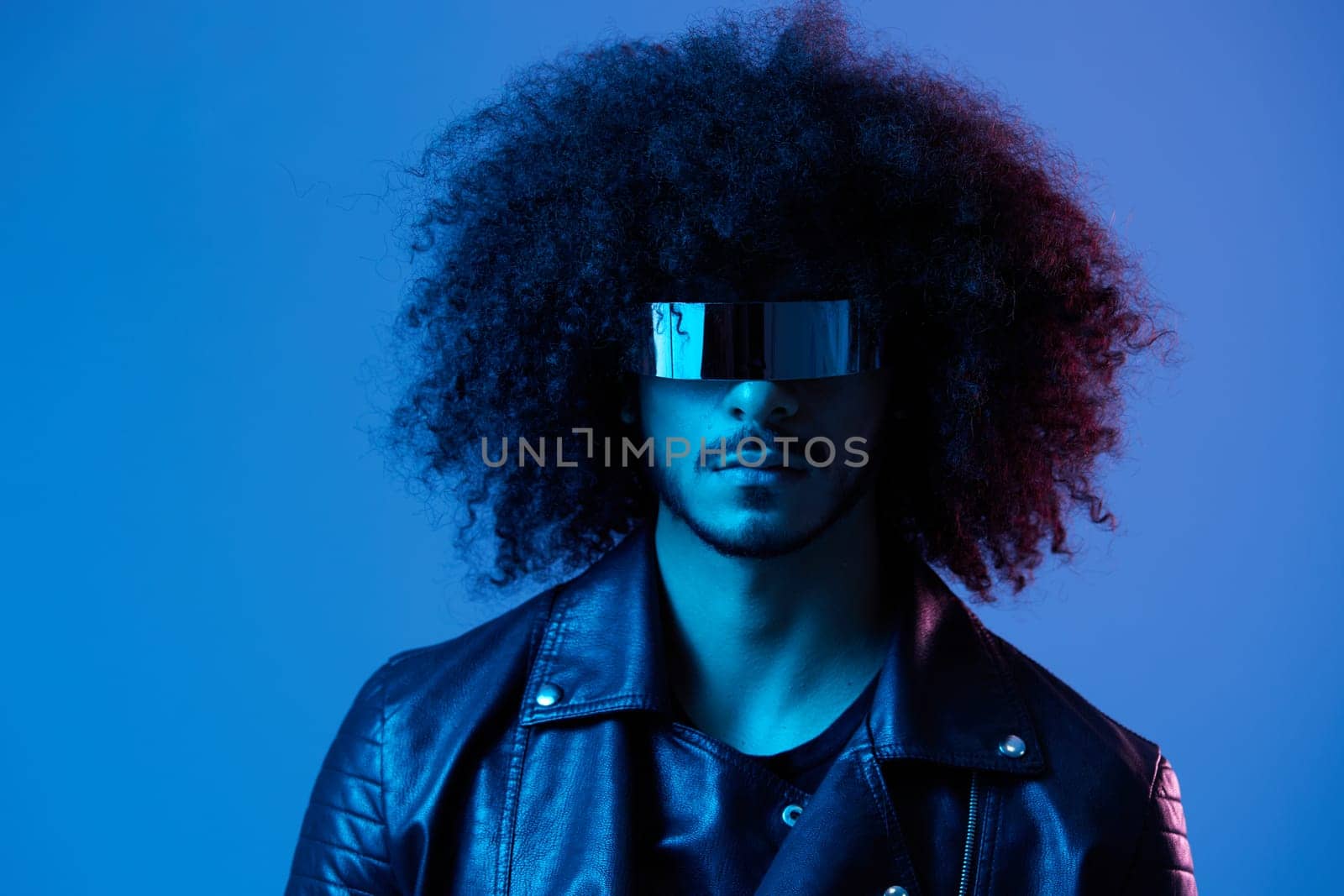 Portrait of fashion man with curly hair with stylish glasses on blue background multinational, colored light, black leather jacket trend, modern concept. by SHOTPRIME