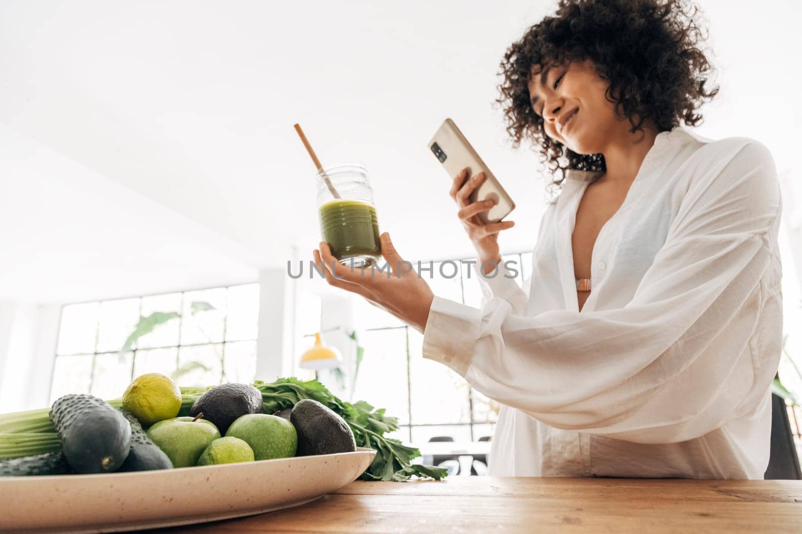 Young african american woman taking photo of green juice and vegetables with cellphone at home. Home concept. Technology concept. Health concept. Copy space