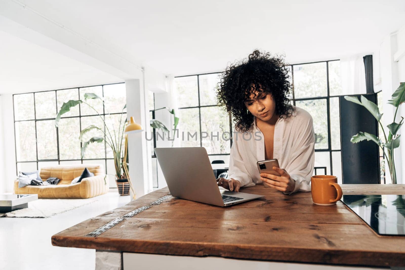 Young african american woman checking cellphone and using laptop in kitchen counter in bright loft apartment by Hoverstock