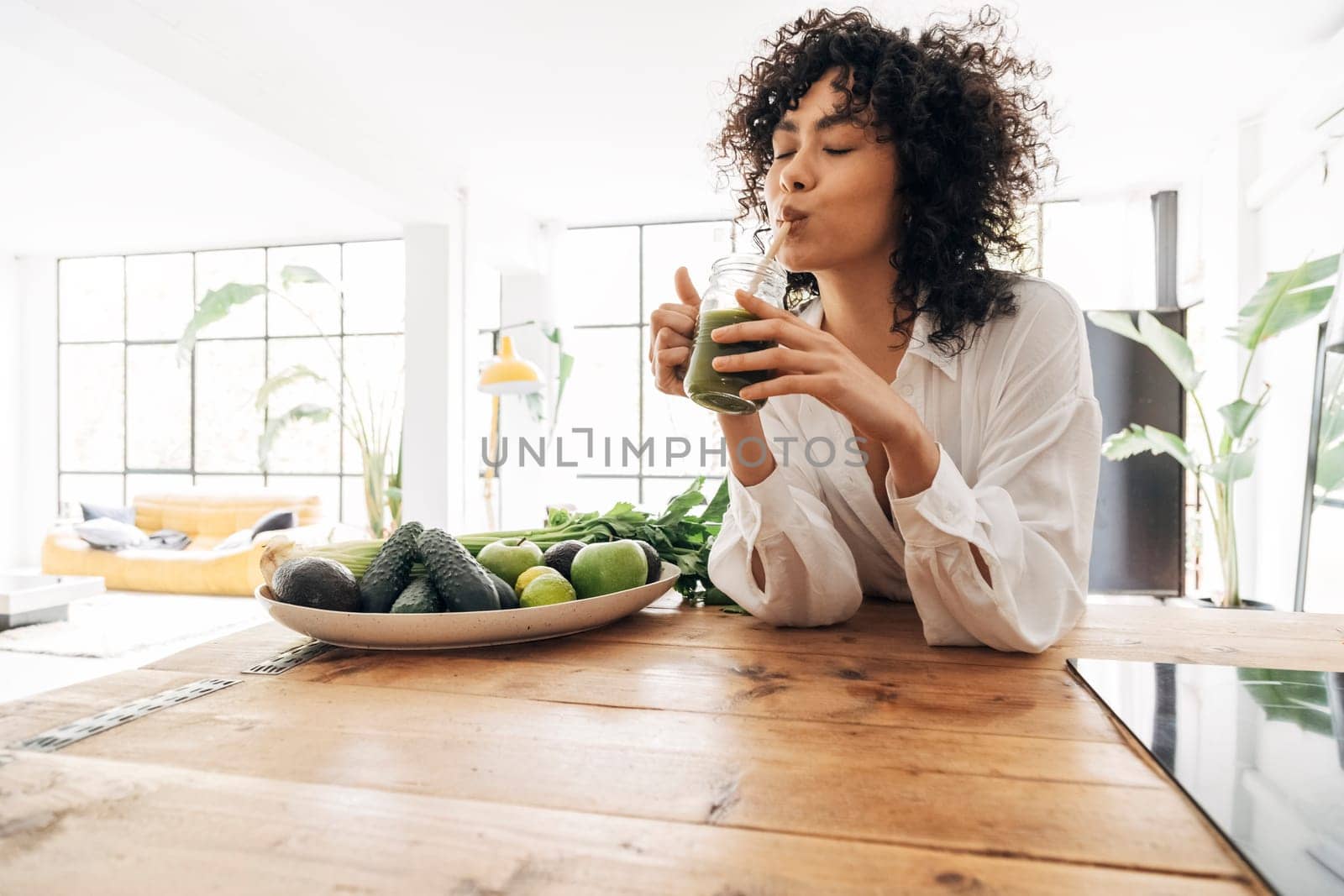 African american woman drinks green juice with bamboo straw in loft apartment by Hoverstock