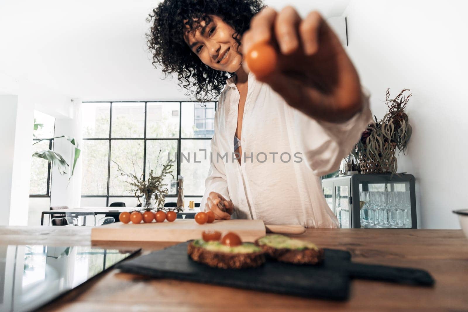 Woman preparing breakfast showing a tomato looking at camera in bright loft kitchen. Selective focus on face. Home concept. Healthy cooking concept. Copy space