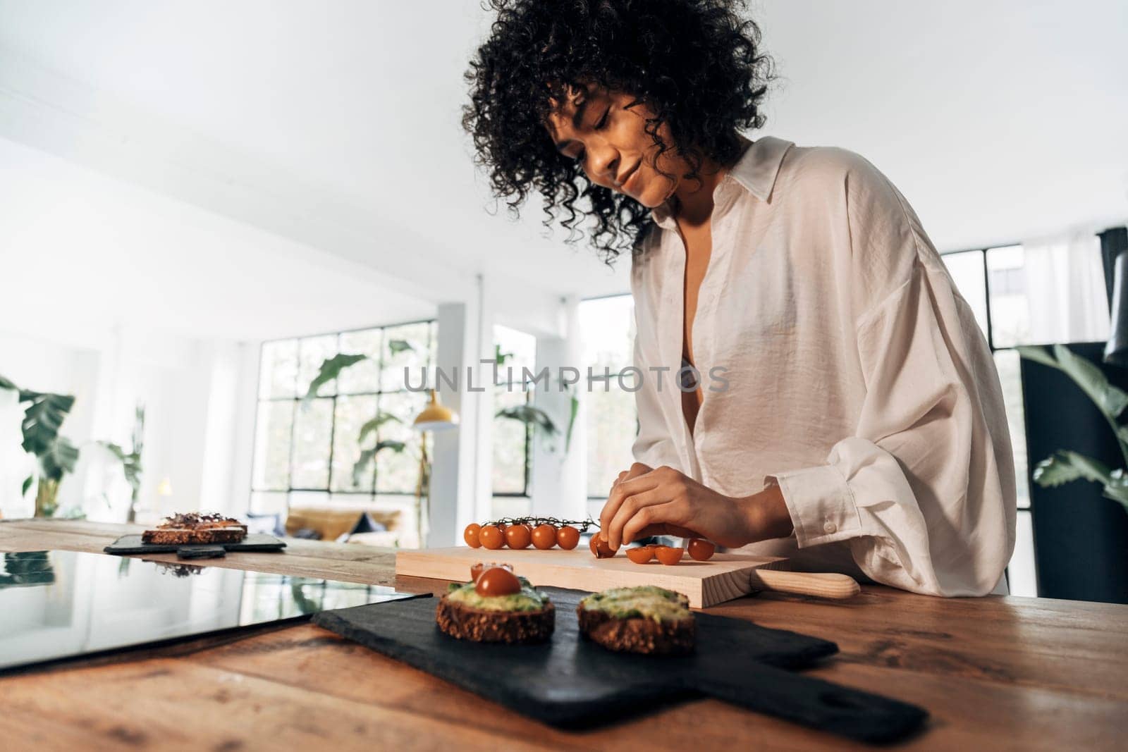 Young african american woman cutting tomatoes preparing breakfast avocado toast in bright loft kitchen by Hoverstock