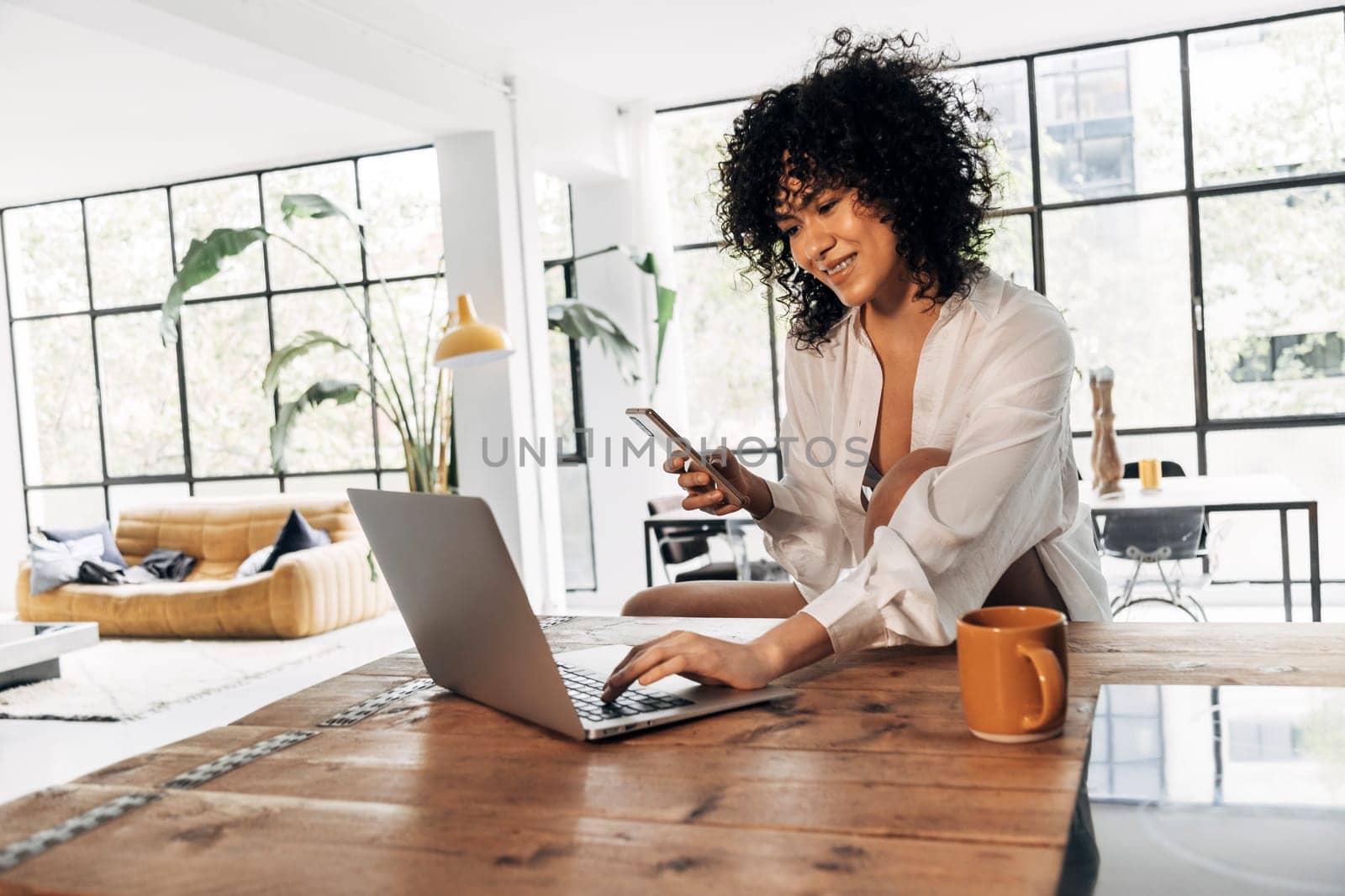 Young beautiful african american woman typing on laptop on kitchen counter, having coffee and holding cellphone by Hoverstock