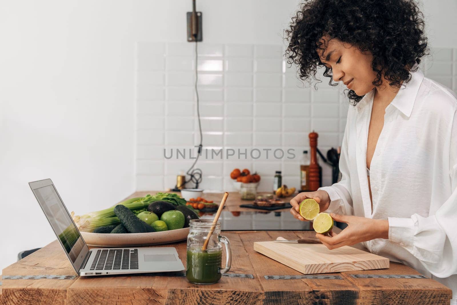 Relaxed Young african american woman preparing green juice for breakfast following online recipe with laptop on kitchen counter. Technology concept. Healthy lifestyle concept. Copy space