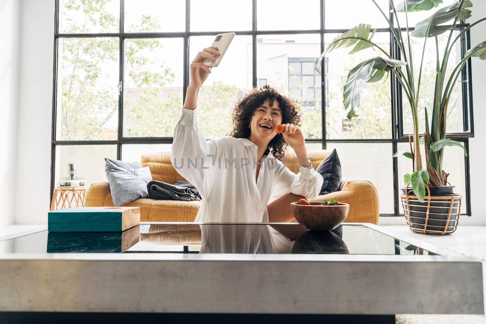 Happy young African American female taking selfie eating a strawberry in big bright living room. Home concept. Technology concept.