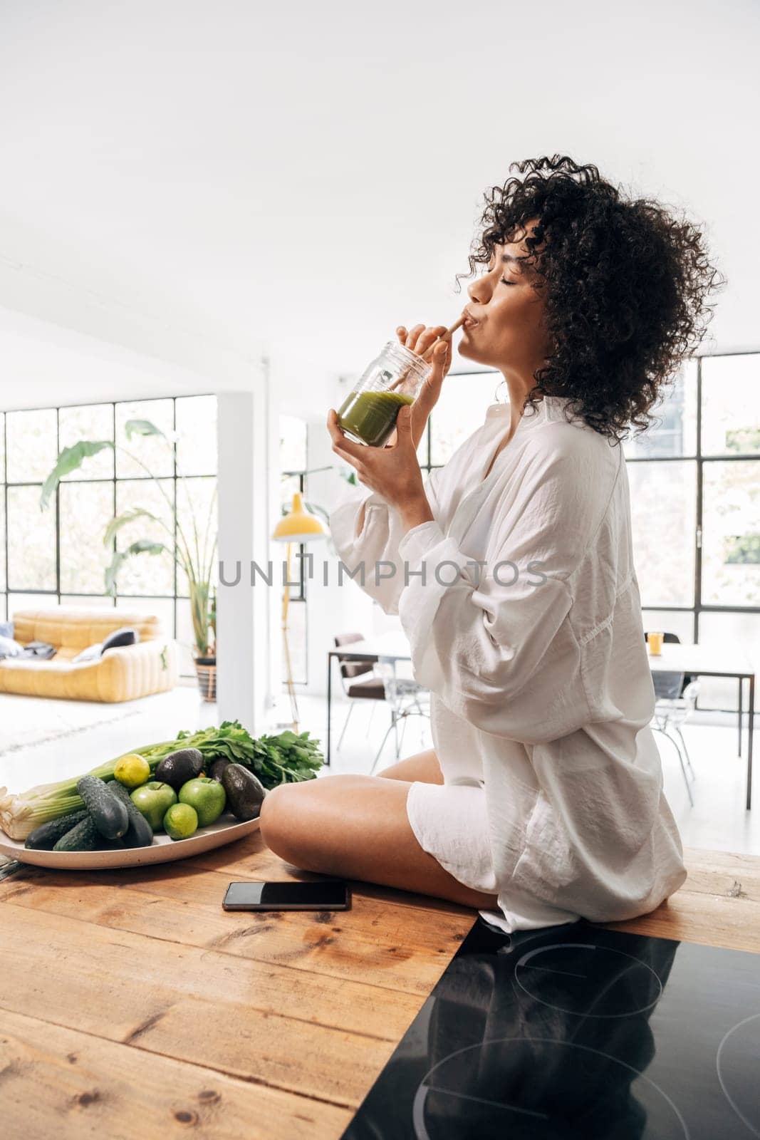 Young african american woman drinking green juice with reusable bamboo straw on kitchen counter. Vertical. by Hoverstock
