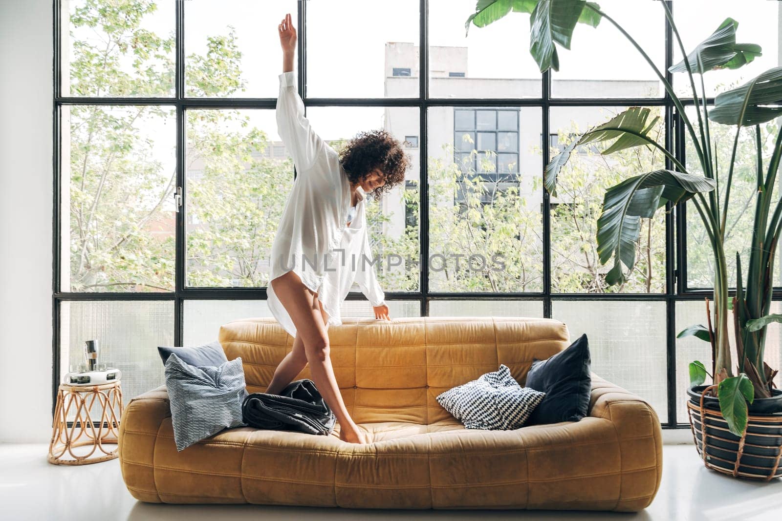 Beautiful African american young woman dancing on couch, having fun, carefree in a big, bright loft apartment by Hoverstock