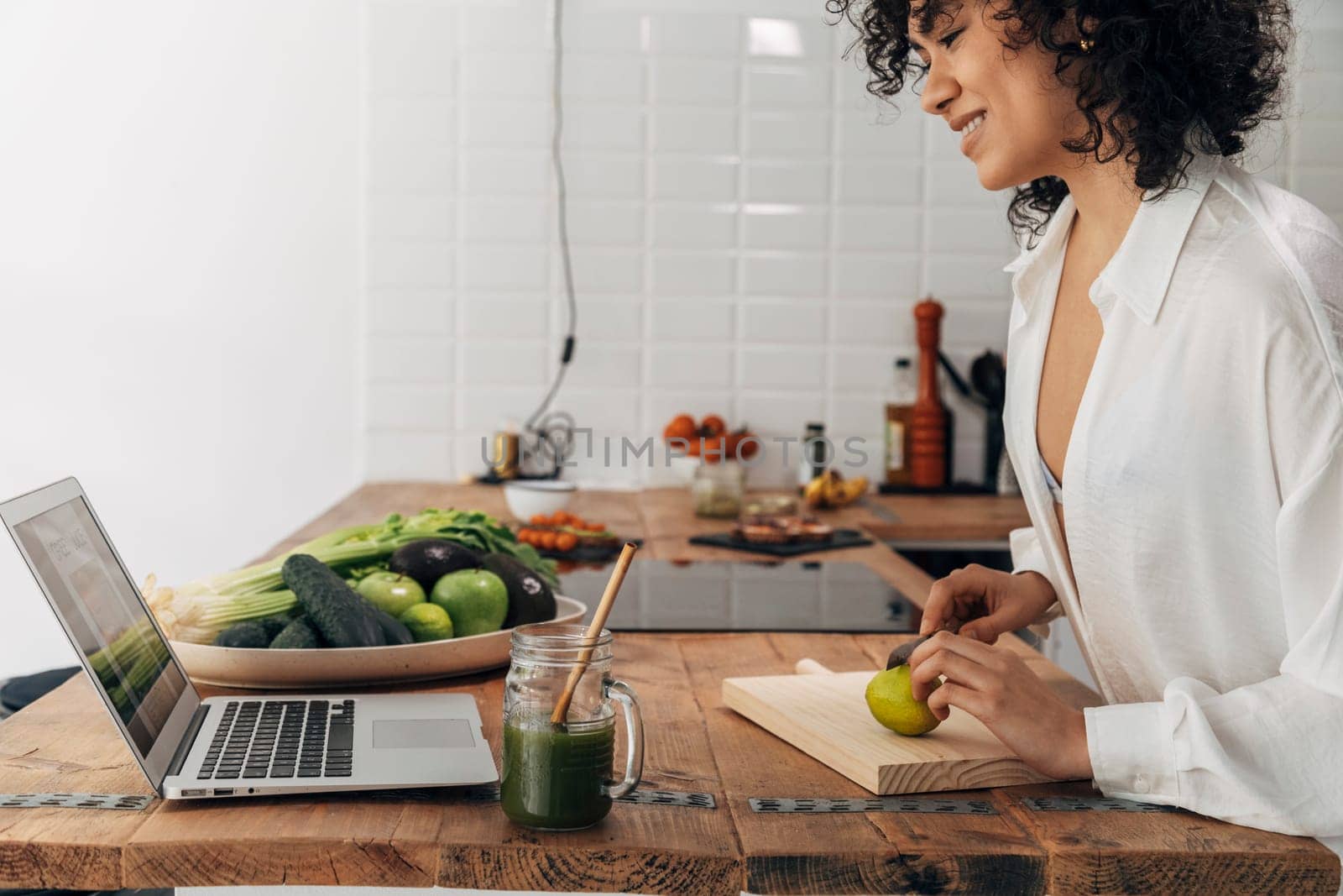 Smiling Young mixed race woman looking recipe laptop. Cutting lime with knife. Kitchen counter top. Green juice reusable straw. Technology concept. Health concept. Home concept. Copy space