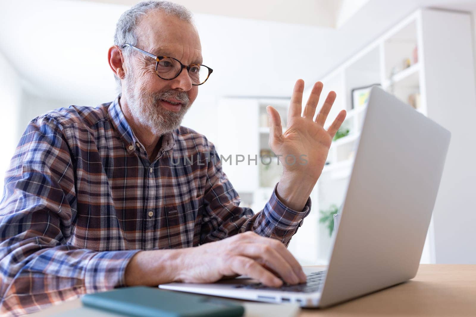 Senior Caucasian man waving hand during video call at home using laptop. by Hoverstock