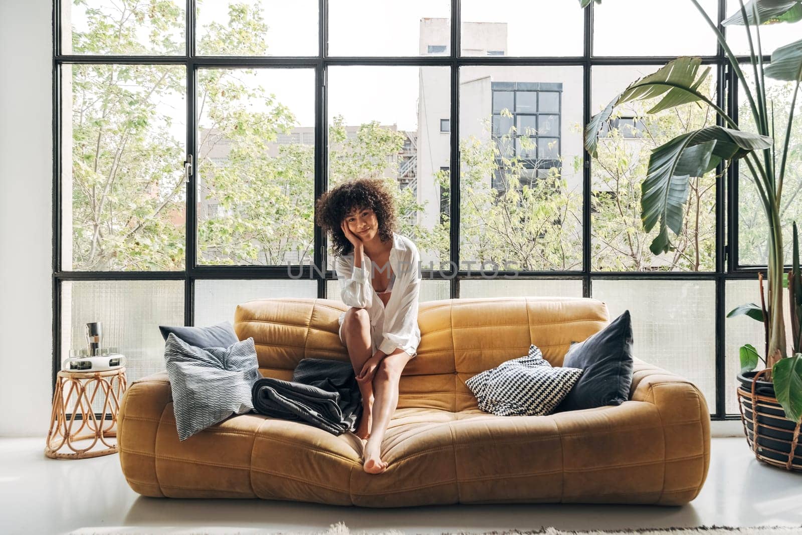 Young and beautiful african american woman with curly hair sitting on a yellow couch looking at camera in a bright loft apartment. Copy space.