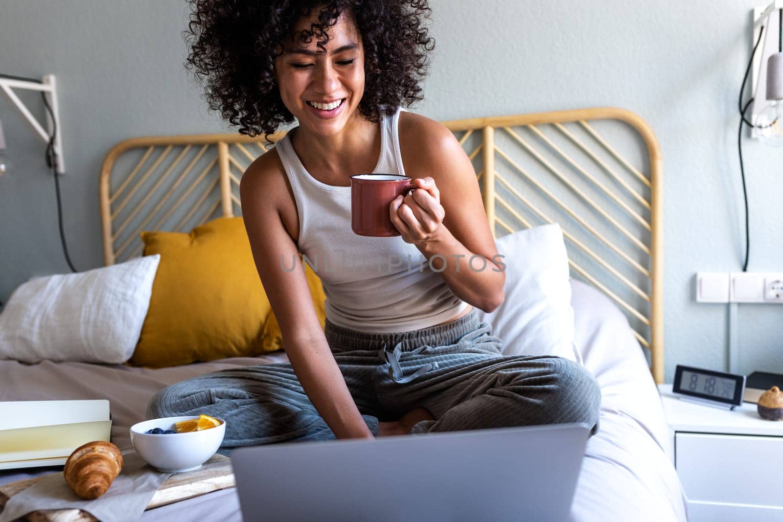 Happy young multiracial latina woman sitting on bed typing on laptop while having morning coffee and eating breakfast. by Hoverstock