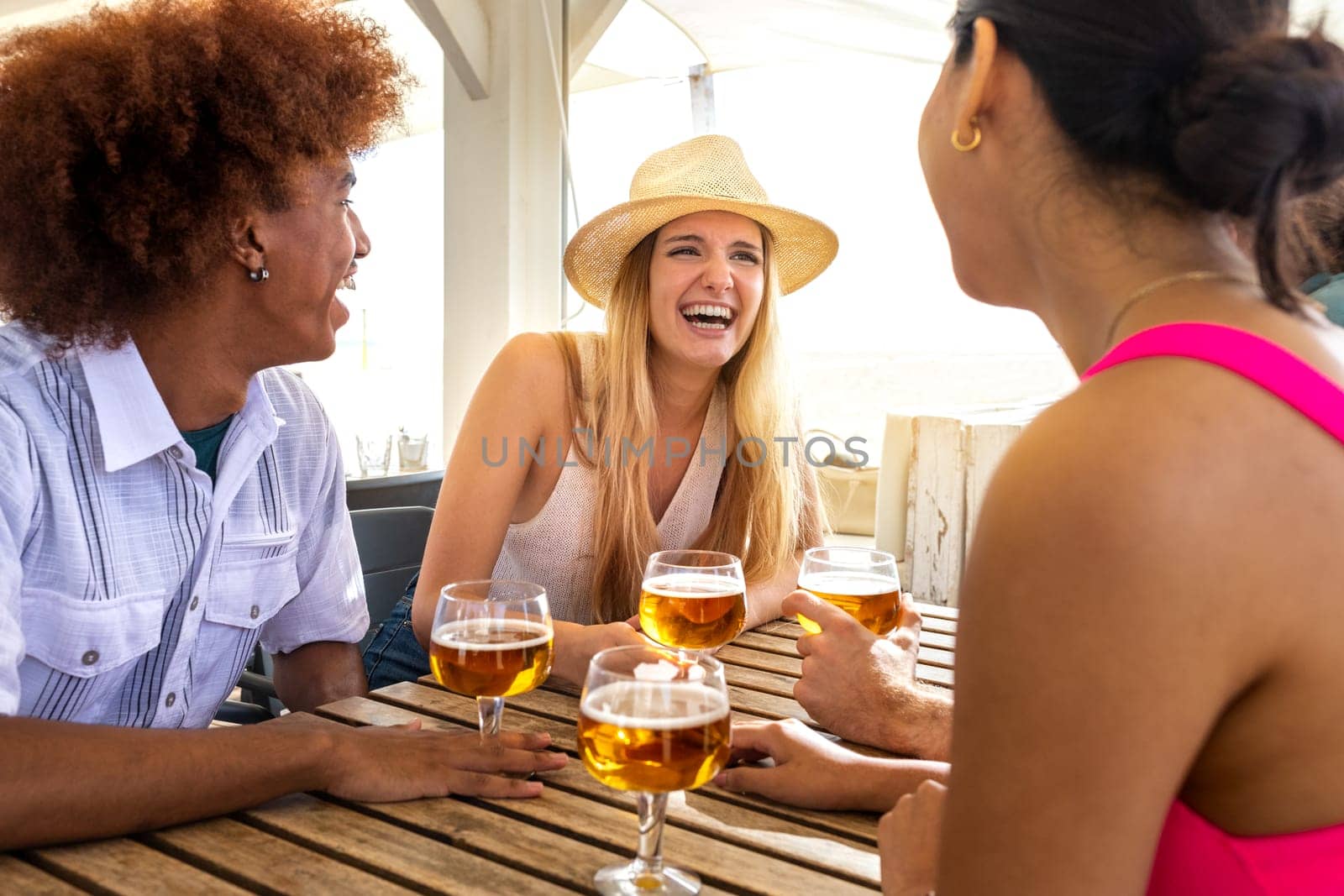 Young caucasian blond woman laughing and enjoying vacation with friends. Multiracial people having beer together at a beach bar. Lifestyle concept.