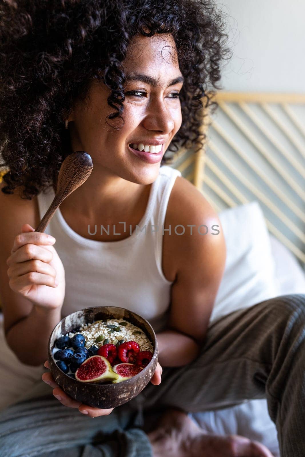 Vertical portrait of happy young latina woman smiling eating healthy breakfast bowl of oats and fruit sitting on bed. by Hoverstock