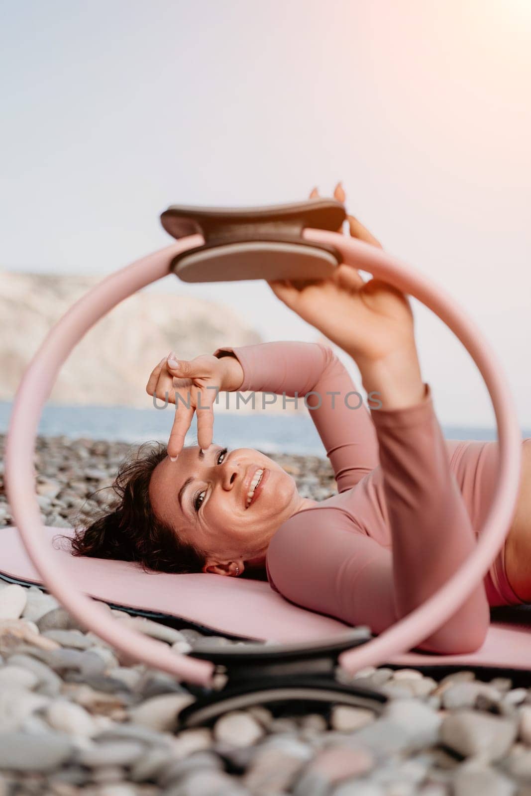 Middle aged well looking woman with black hair doing Pilates with the ring on the yoga mat near the sea on the pebble beach. Female fitness yoga concept. Healthy lifestyle, harmony and meditation.