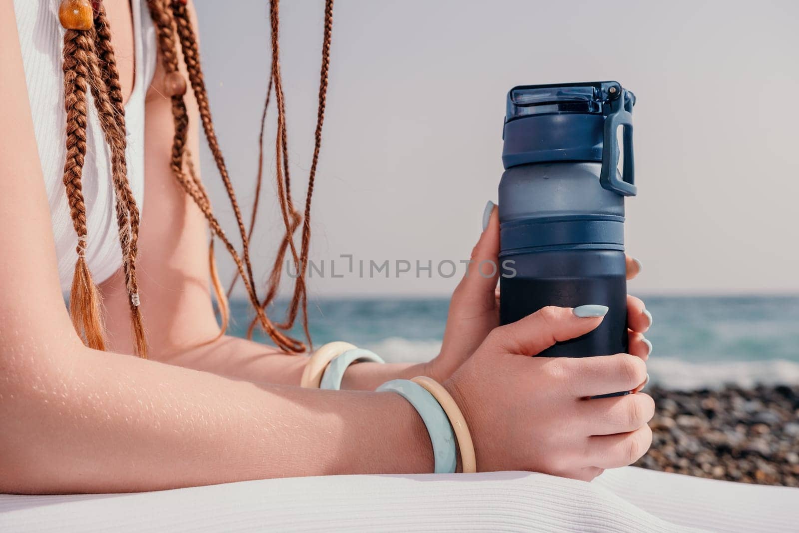 Woman sea yoga. Thermo mug in the hands of woman with braids dreadlocks in white leggings and tops doing stretching pilates on yoga mat near sea. Female fitness yoga routine concept. Healthy lifestyle by panophotograph