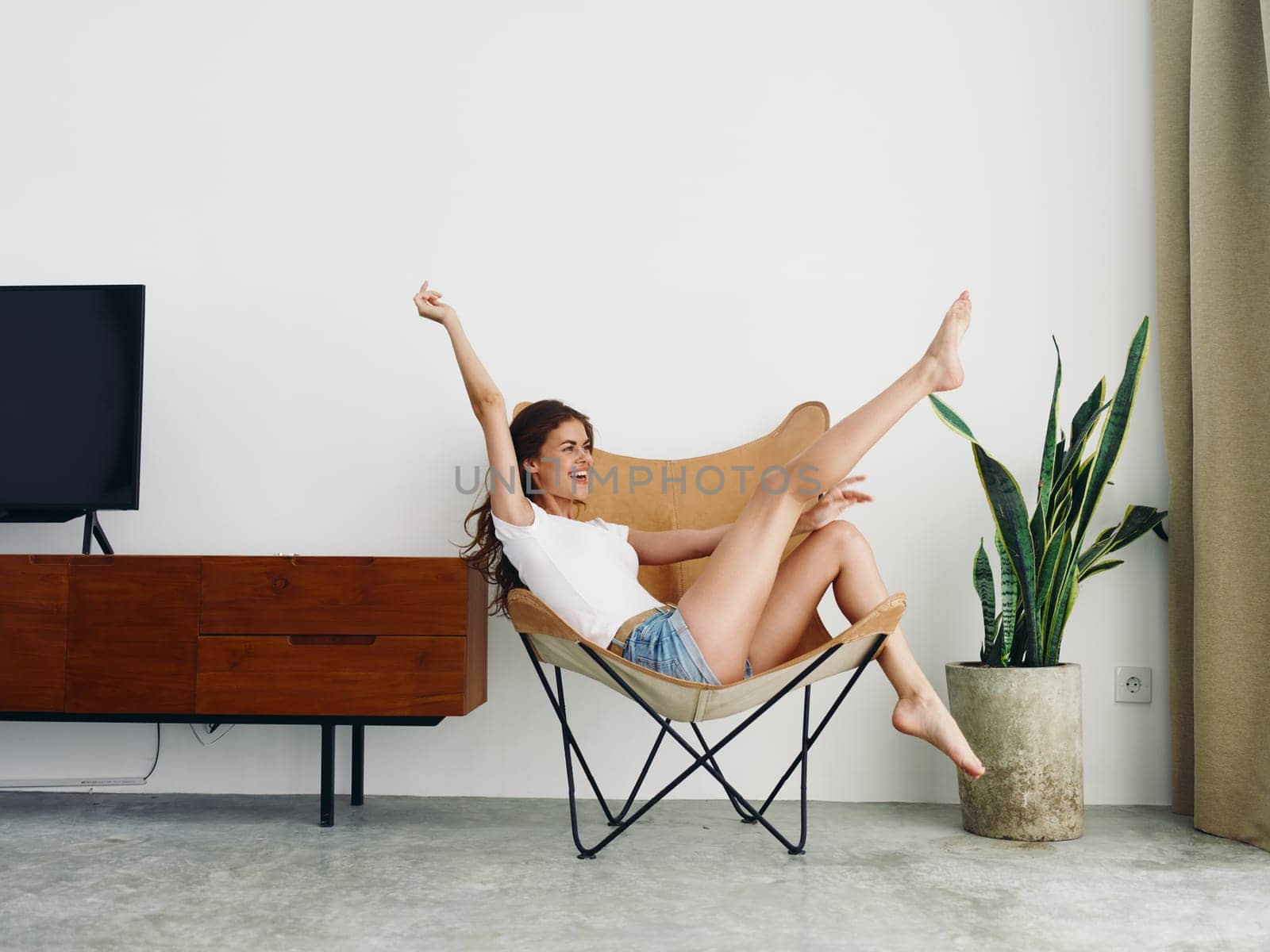 Woman lying in a leather armchair raised arms and legs cheerful happiness and a smile, relaxing at home stylish modern interior with white walls, copy space. by SHOTPRIME