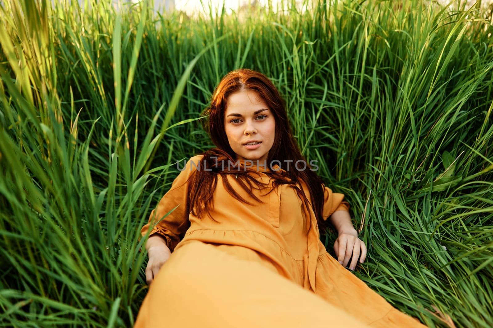 a happy woman lies in the tall green grass in a long orange dress and smiling pleasantly looks into the camera enjoying nature. Close horizontal photo. High quality photo
