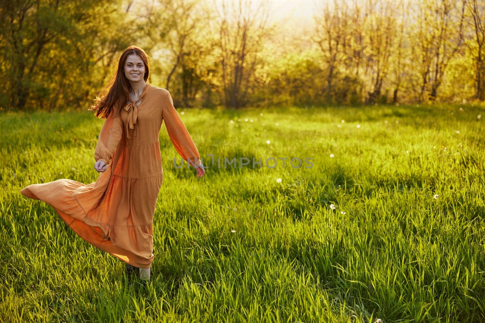 a joyful woman stands green in a field smiling at the camera, in a long orange dress, illuminated by the warm rays of the setting sun by Vichizh