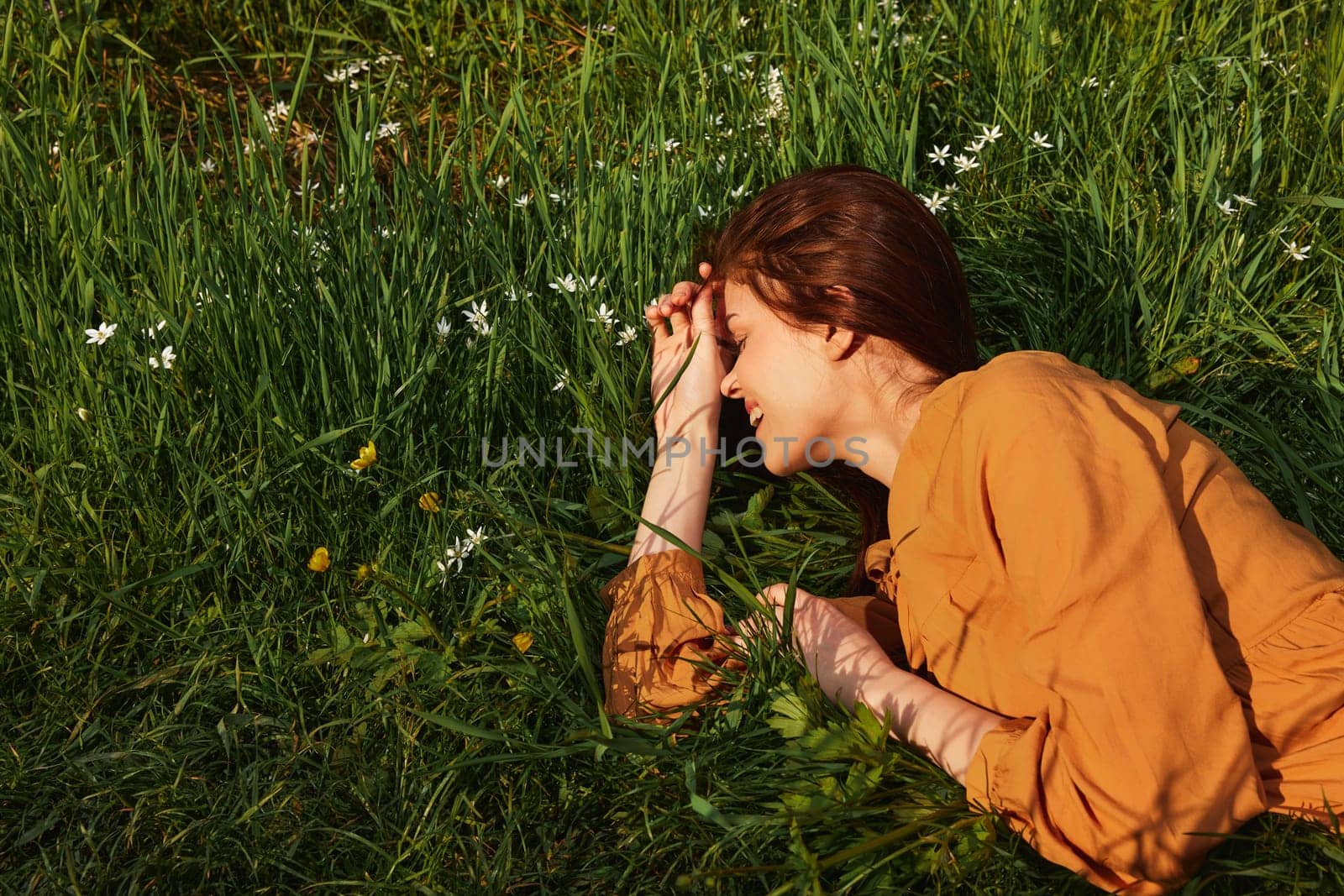 a calm woman with long red hair lies in a green field with yellow flowers, in an orange dress, smiling pleasantly, closing her eyes from the bright summer sun, resting her head on her hands. High quality photo