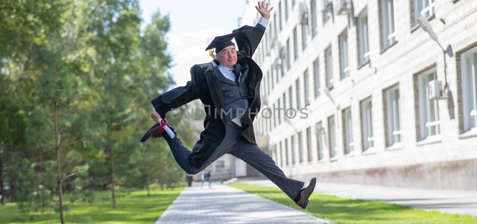 Old happy man in graduation gown jumping outdoors and holding diploma