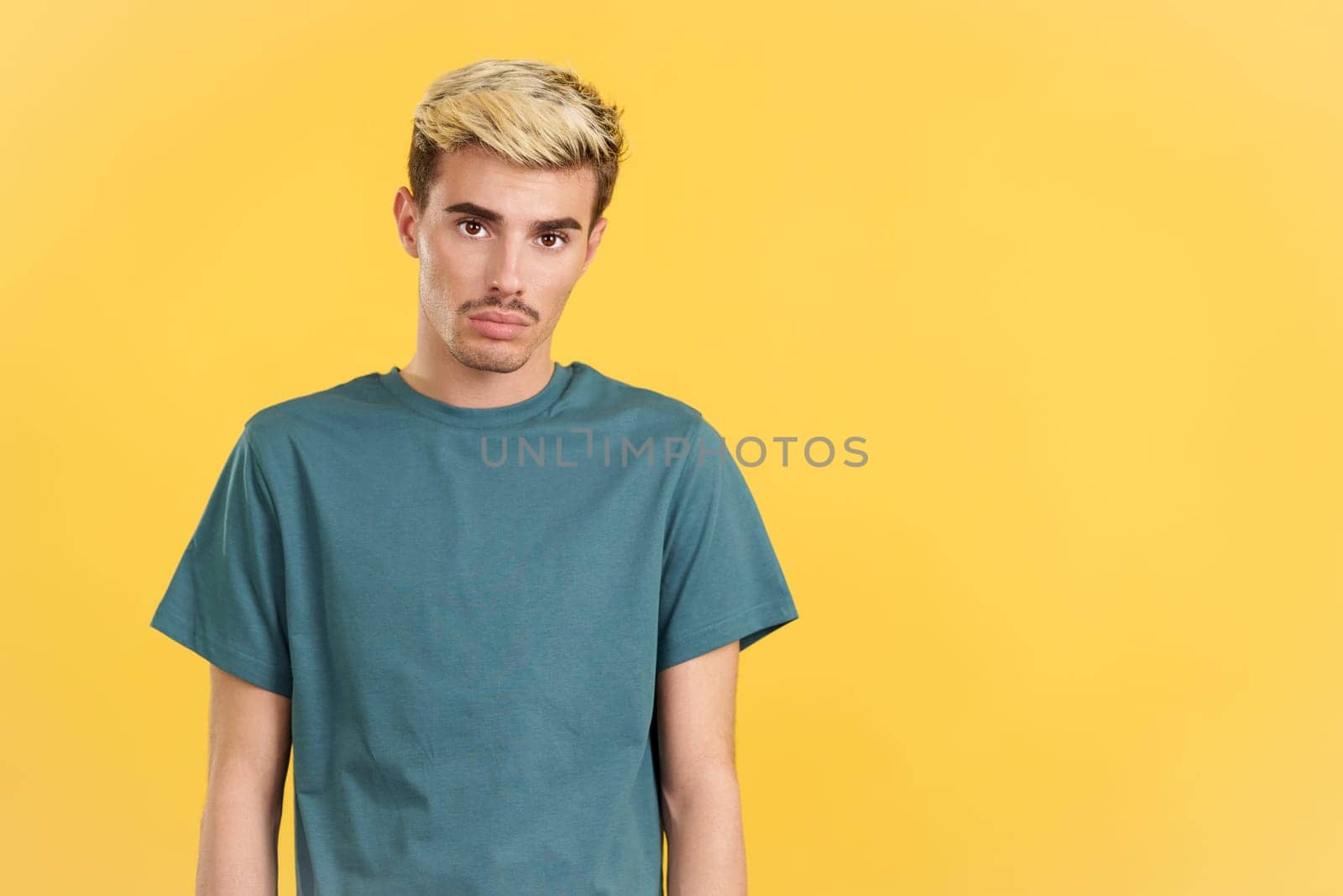 Sad gay man standing and looking at camera in studio with yellow background