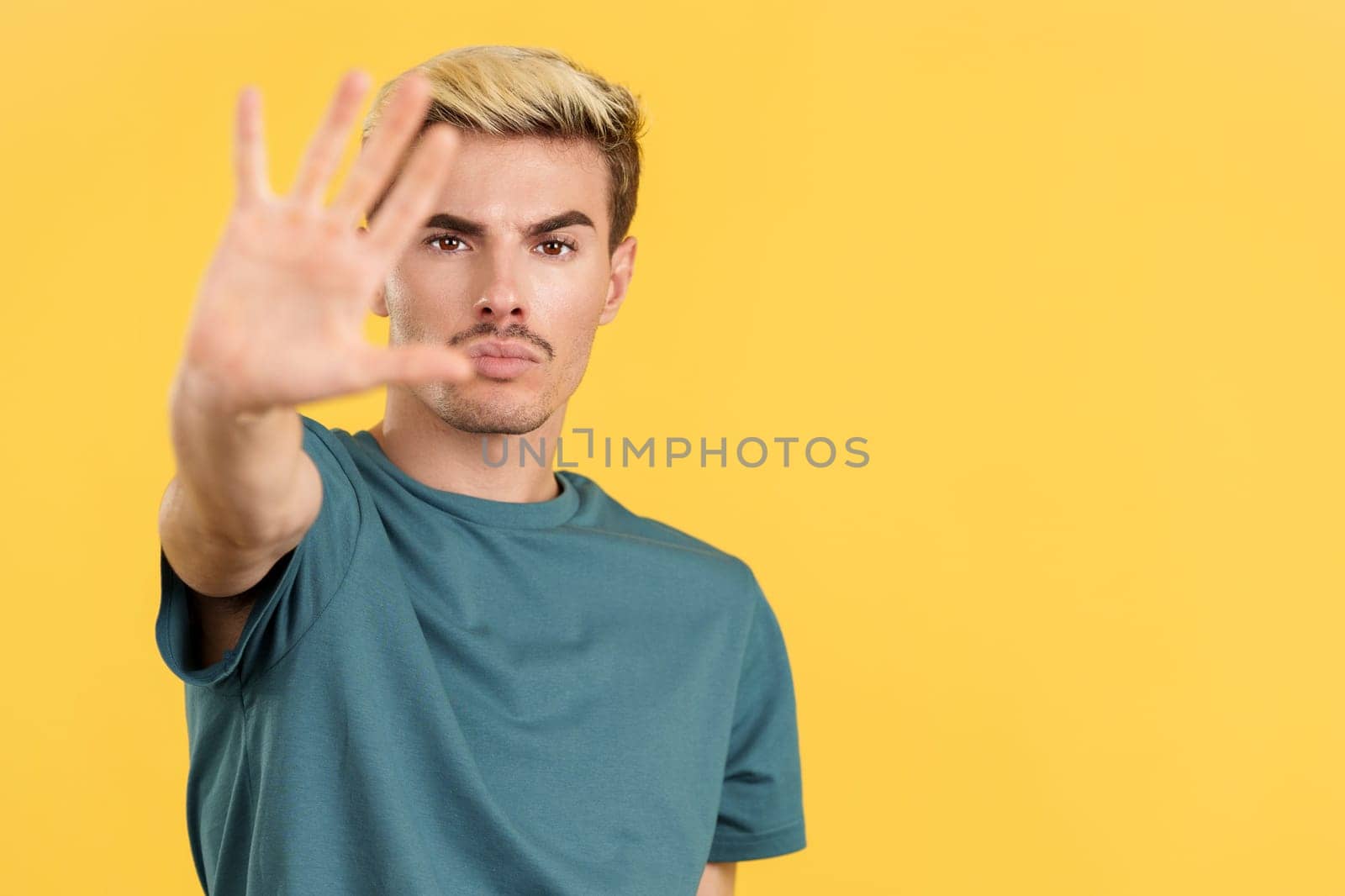 Gay man gesturing prohibition and stop with hand in studio with yellow background