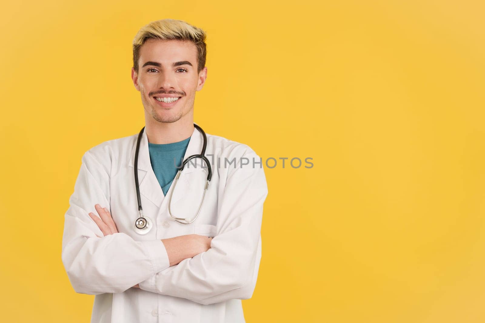 Gay doctor smiling at the camera standing with arms crossed in studio with yellow background