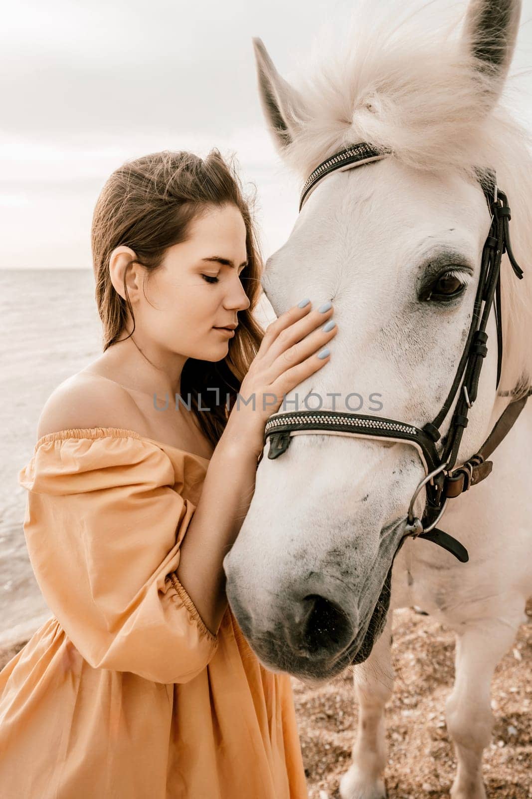A woman in a dress stands next to a white horse on a beach, with the blue sky and sea in the background