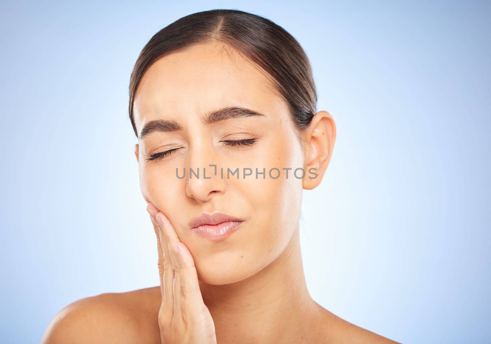 Toothache, pain and cavity with a model woman holding her mouth in studio on a blue background for oral care. Dental hygiene, plaque and face with an attractive young female rubbing her sore cheek.