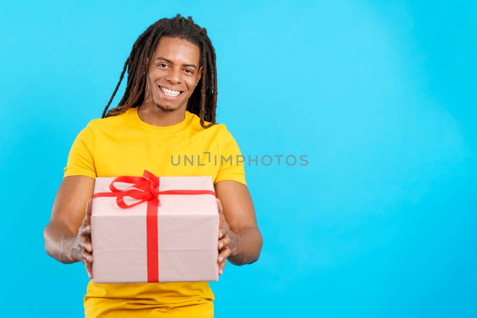 Happy latin man with dreadlocks looking at the camera giving a present in studio with blue background