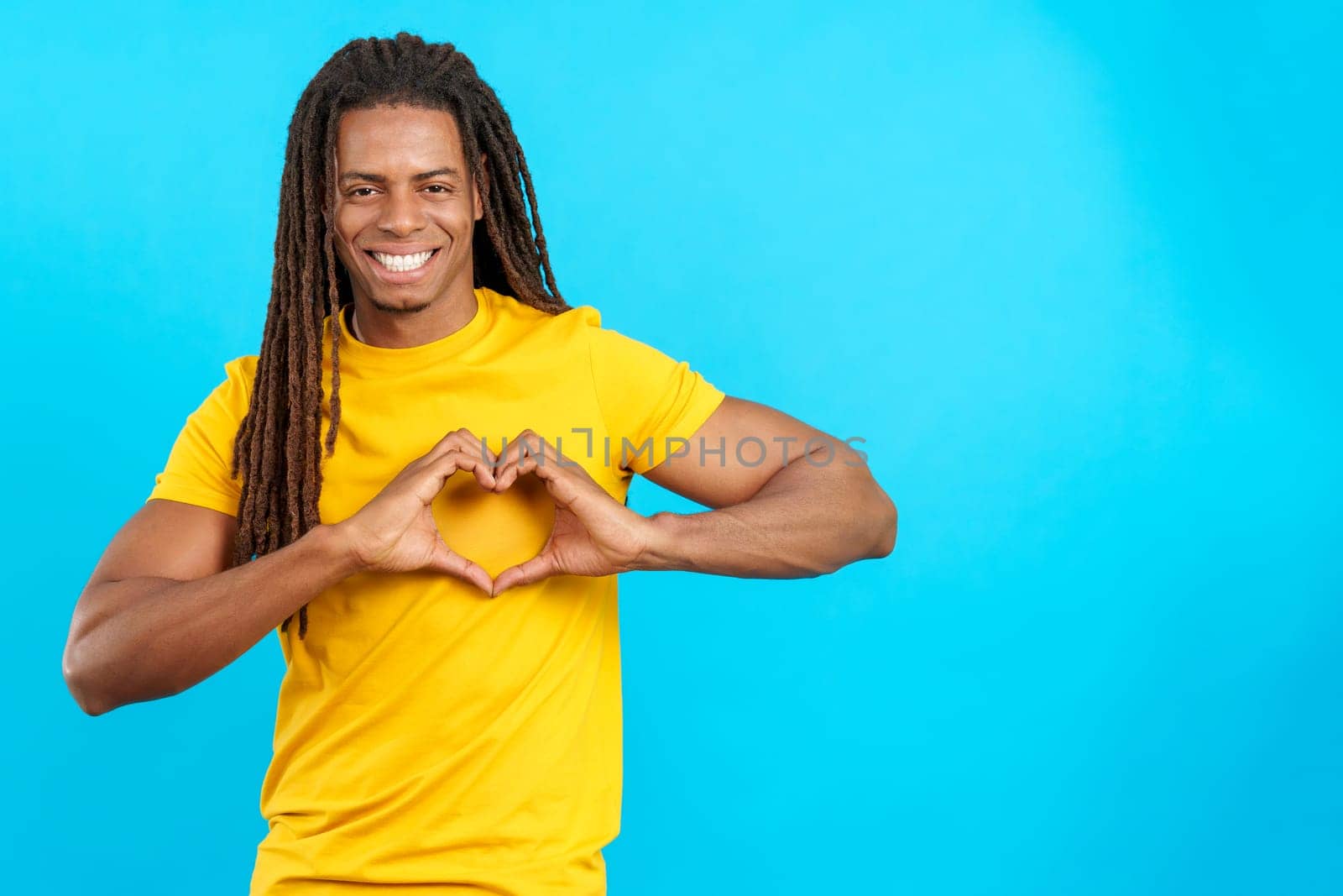 Latin man with dreadlocks representing a heart in the shape with hands in studio with blue background