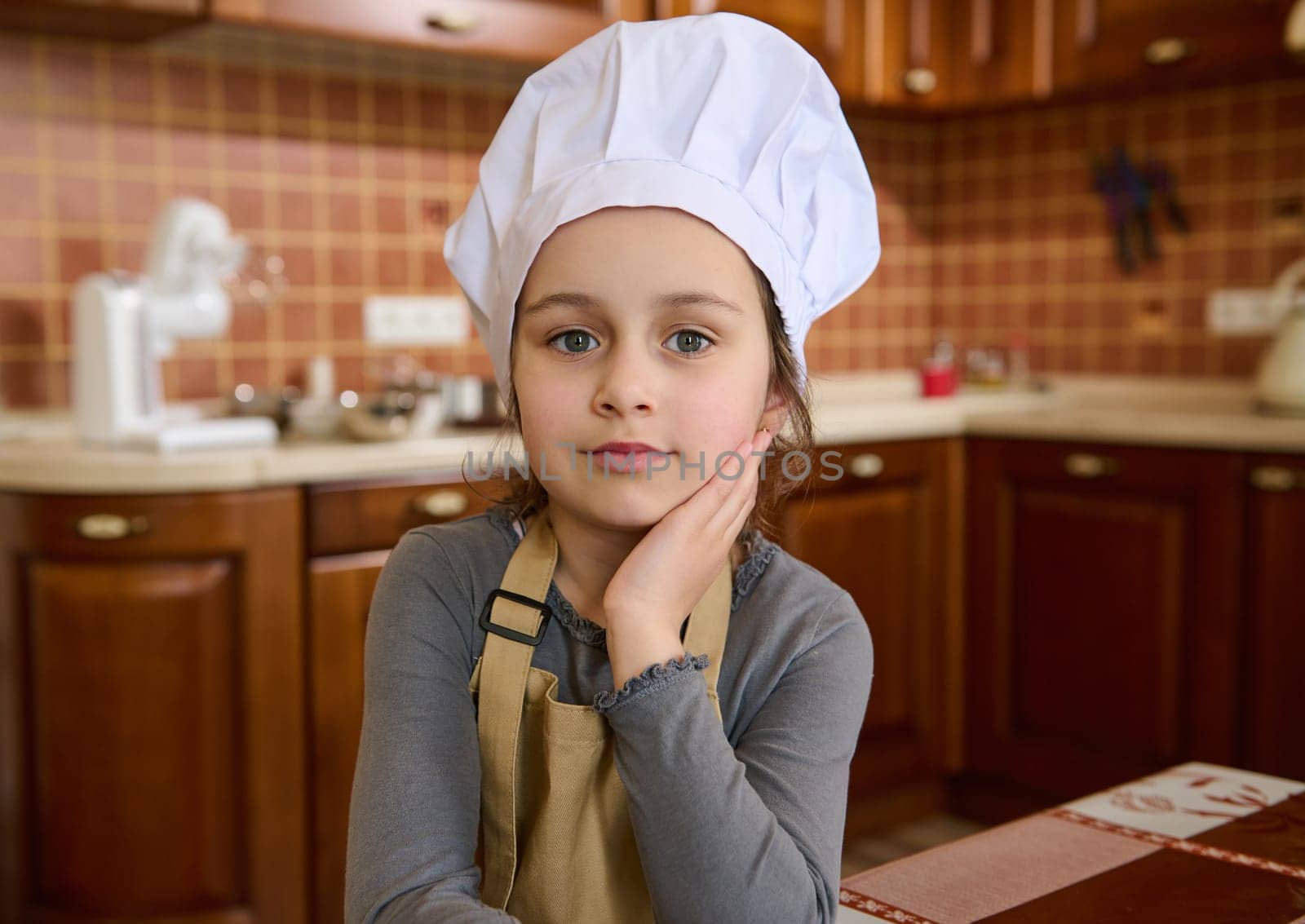 Authentic portrait of Caucasian adorable child girl in white chef hat and beige apron, little baker confectioner looks at camera, standing at home kitchen interior. Cooking class. Kids learn culinary