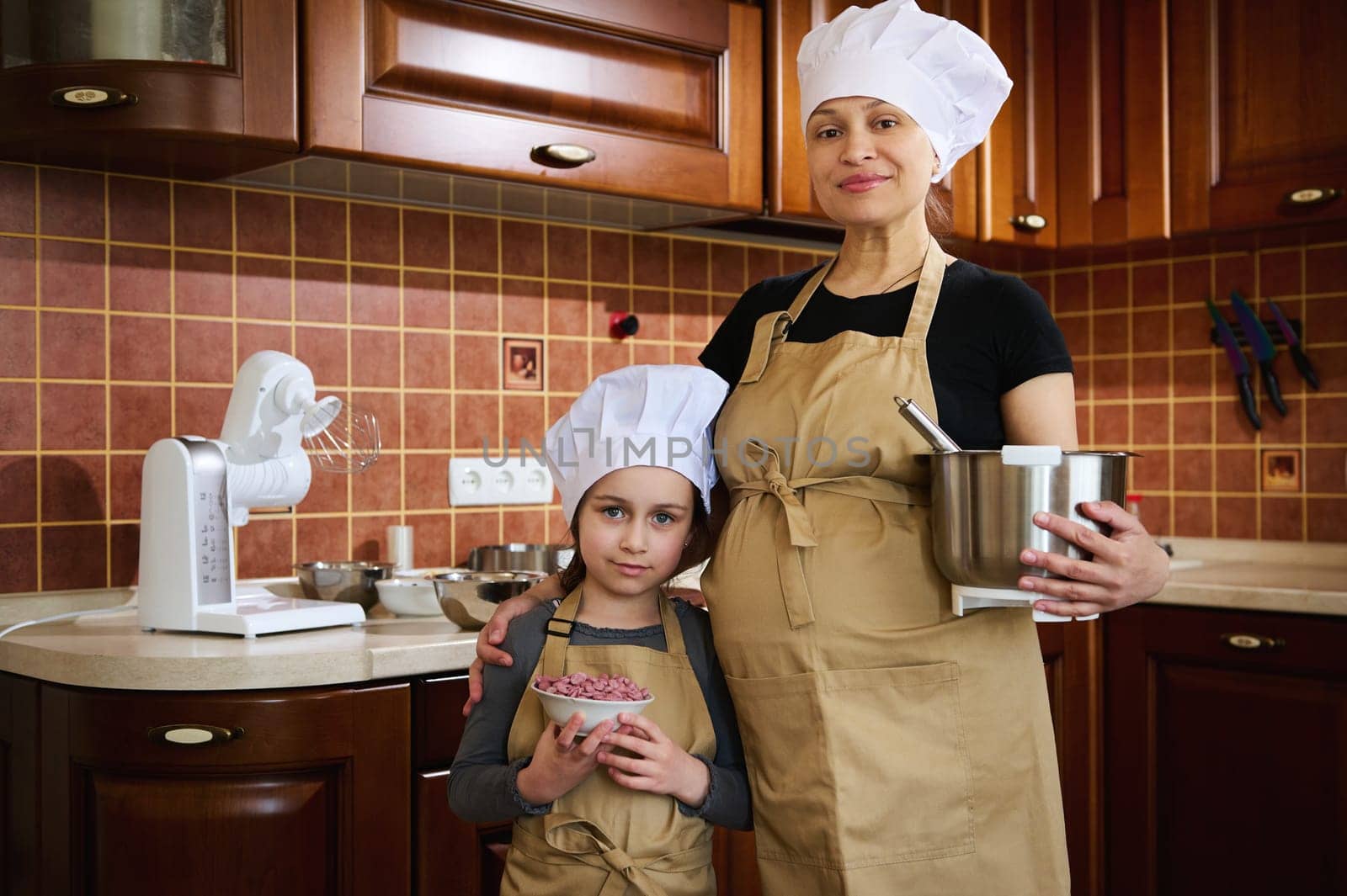 Happy pregnant woman, expectant mother and her lovely little daughter, dressed in apron and chef's hat, enjoying cooking together, standing in the home kitchen, smiling, confidently looking at camera