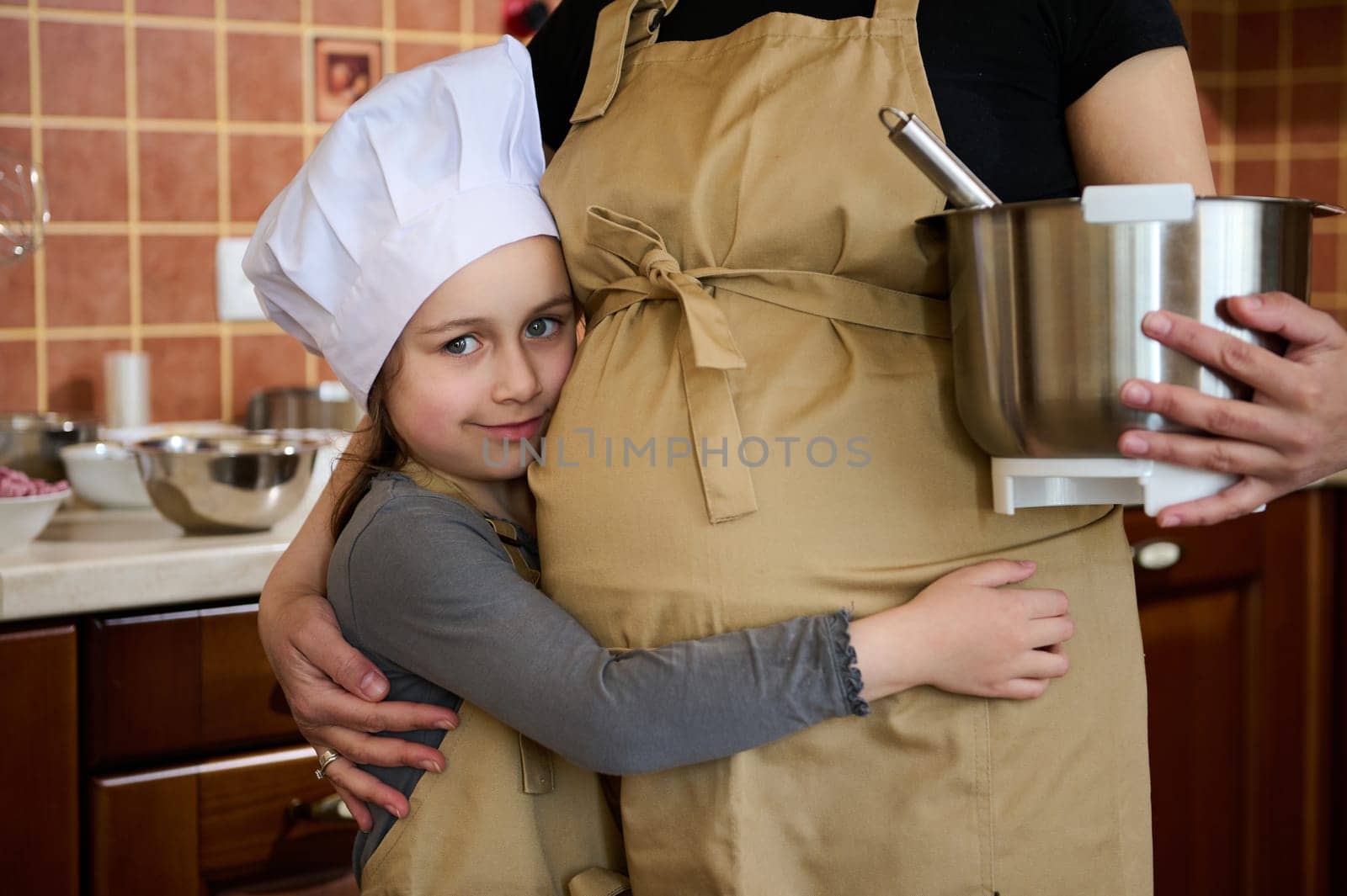 Adorable little girl gently hugging the pregnant belly of her mother, standing in the kitchen while preparing delicious homemade dessert, looking at camera. Mom and daughter cooking together. Family