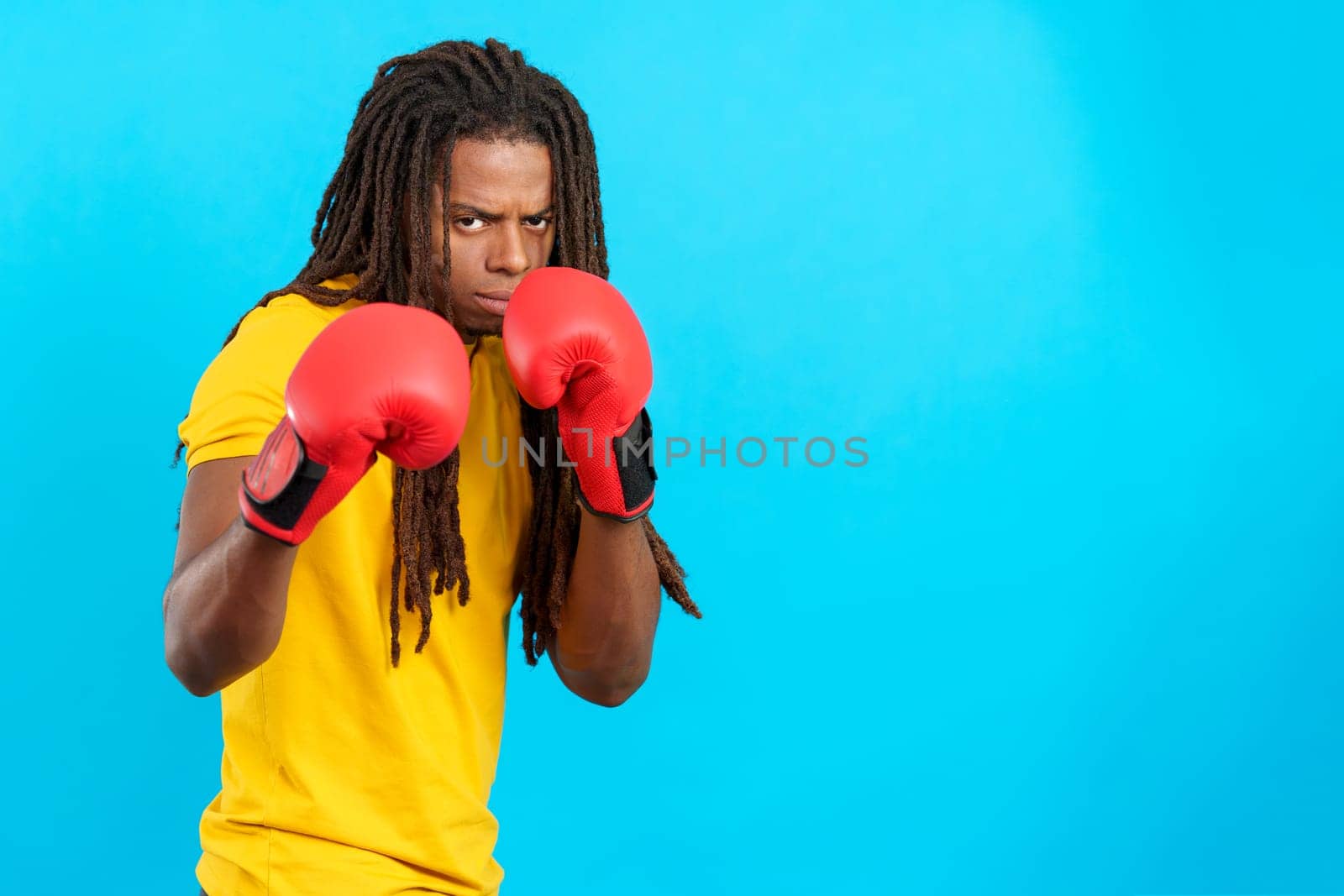 Latin man with dreadlocks wearing boxing gloves in fight pose in studio with blue background