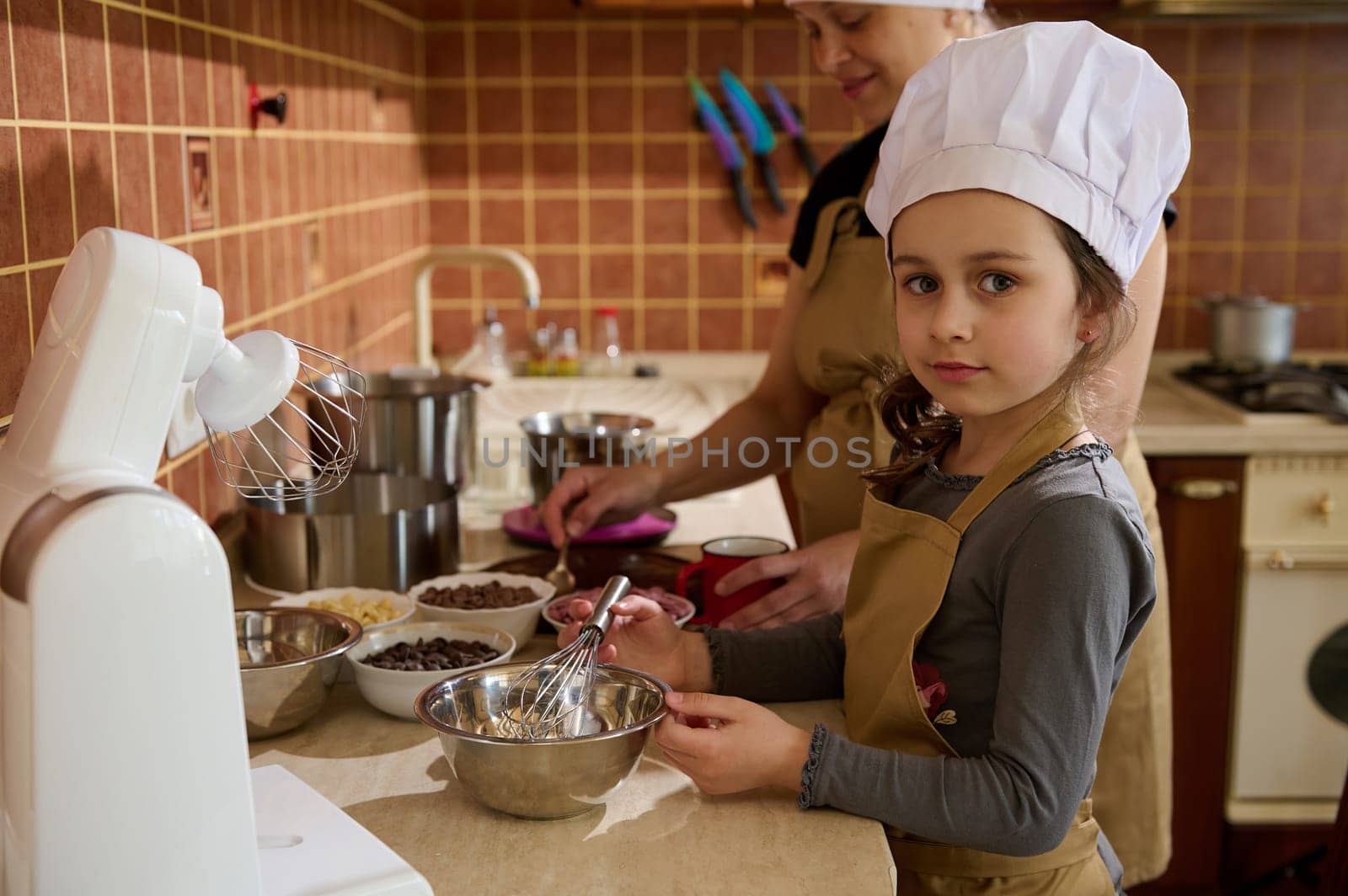 Portrait of a beautiful kid girl in white chef's hat and beige apron standing by kitchen counter, helping her mother, mixing whipped cream with melted chocolate using a whisk, looking at the camera