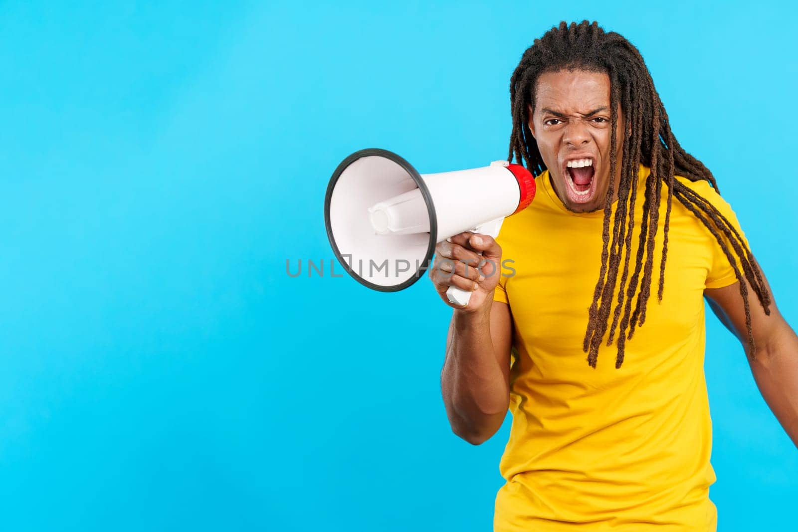 Angry latin man with dreadlocks shouting using a megaphone in studio with blue background