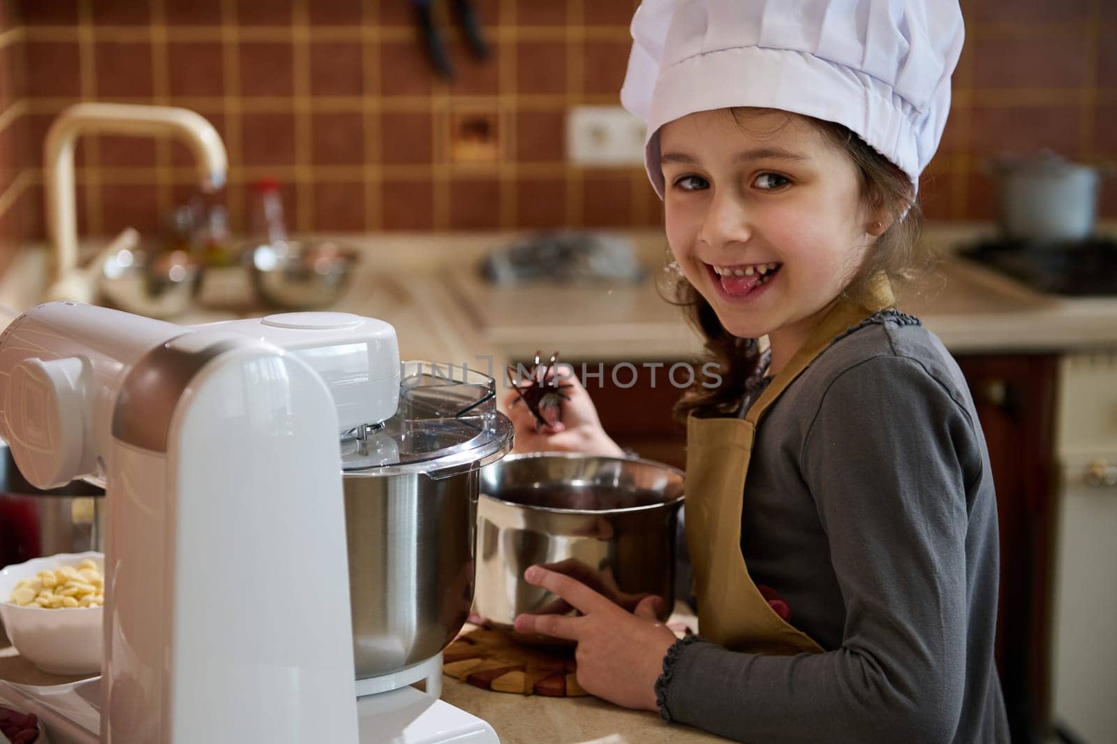 Mischievous child girl 5 years old, tastes melted Belgian chocolate, standing at the kitchen countertop, smiling slyly at the camera. Little kid pastry chef dressed in white chef's hat and beige apron
