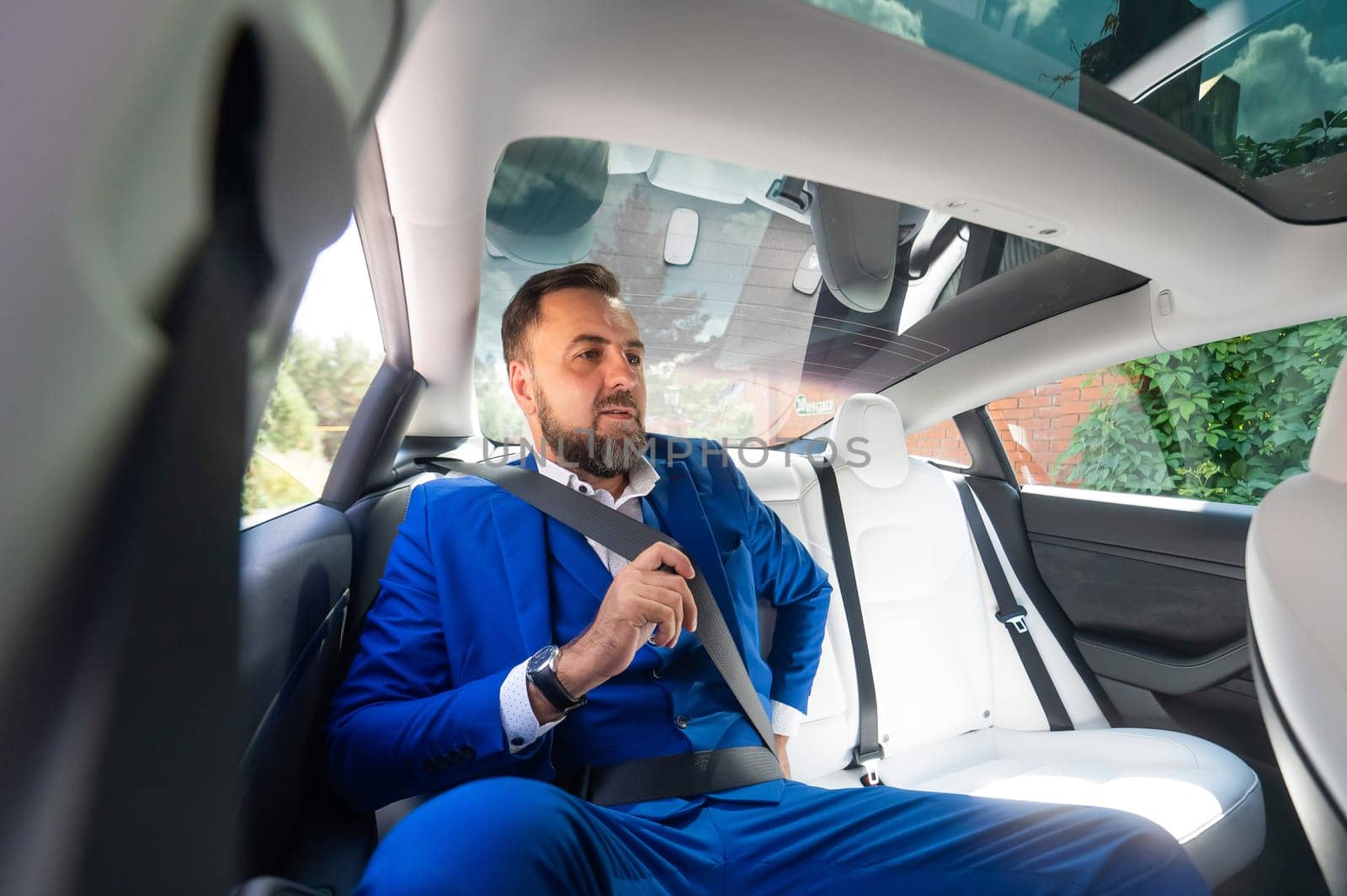 A caucasian man in a blue suit fastens his seat belt in the back seat of a car. Business class passenger