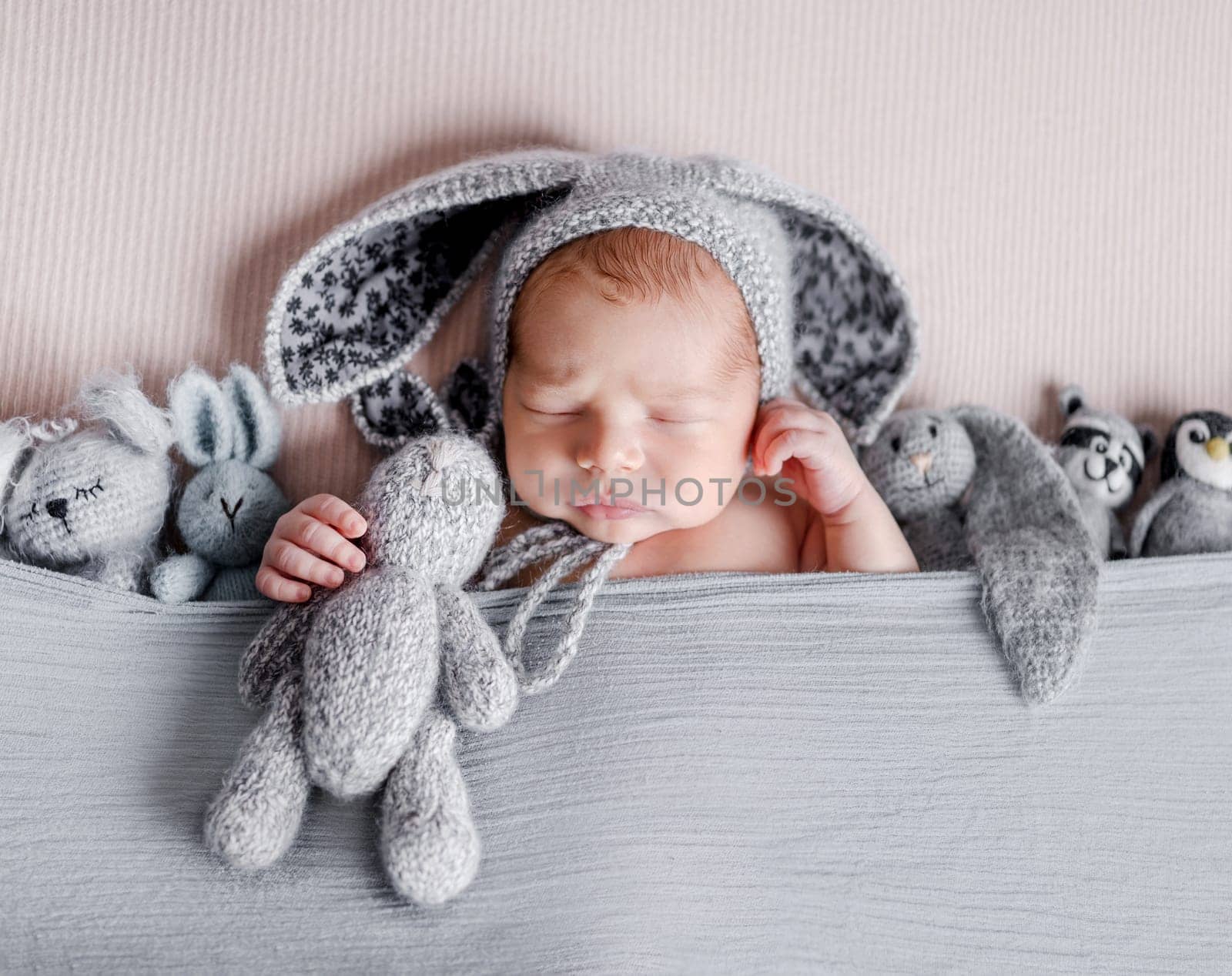 Cute newborn baby boy sleeping wearing bunny ears hat and holding knitted handmade toys. Adorable infant child kid napping studio portrait