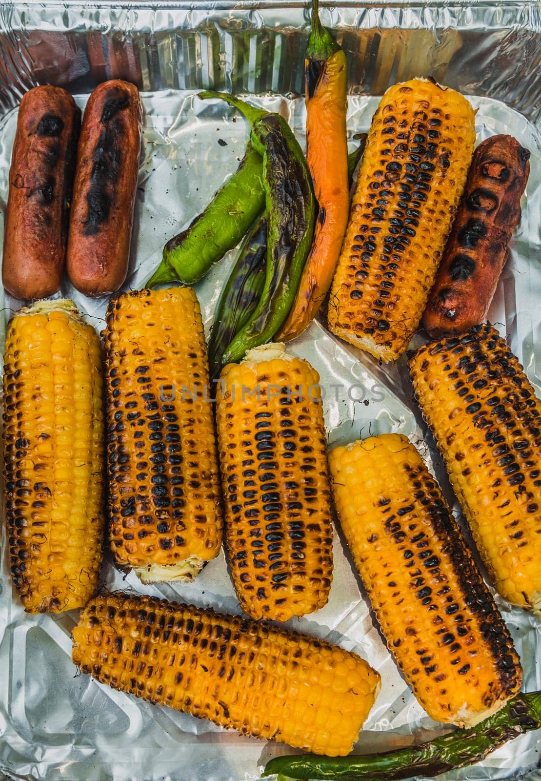 Top view of grilled corn cobs, green chili pepper and sausages on disposable aluminum  food packaging.