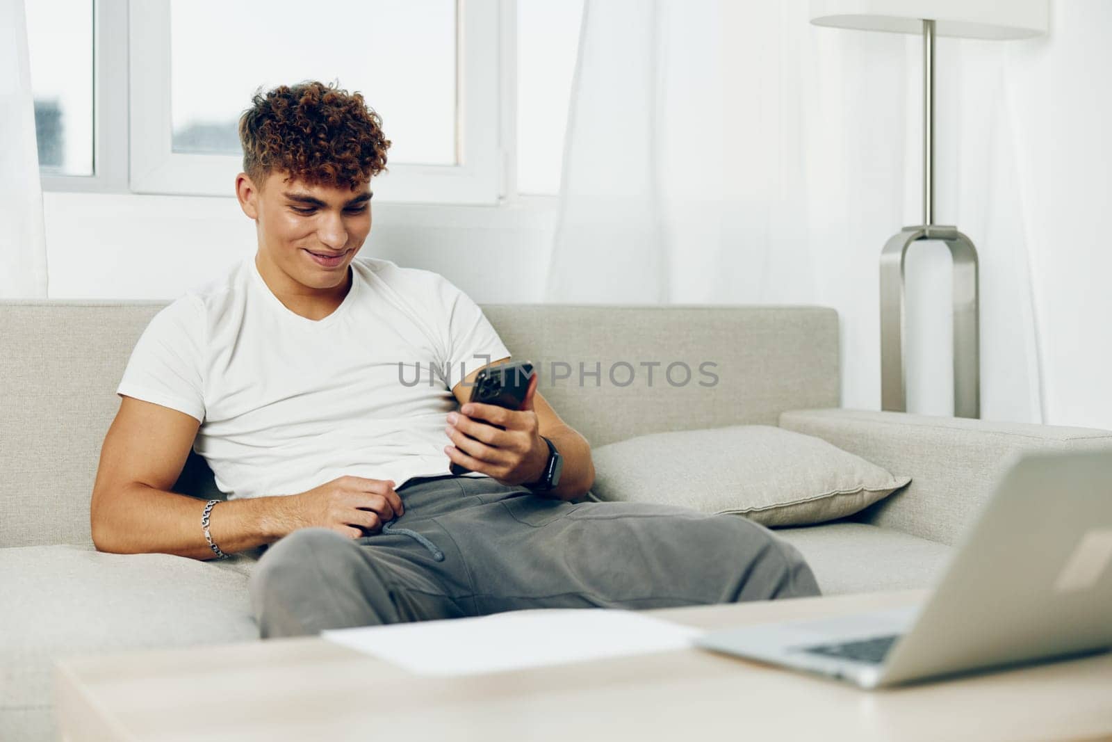 man interior mobile laptop freelancer blissful smile lifestyle couch blogger using adult text message by SHOTPRIME