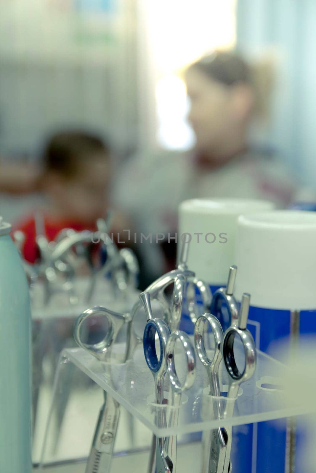 Closeup on hairdressing scissors in professional hairdressing salon. Little boy sit on his mother and get a haircut on blurred mirror reflection.