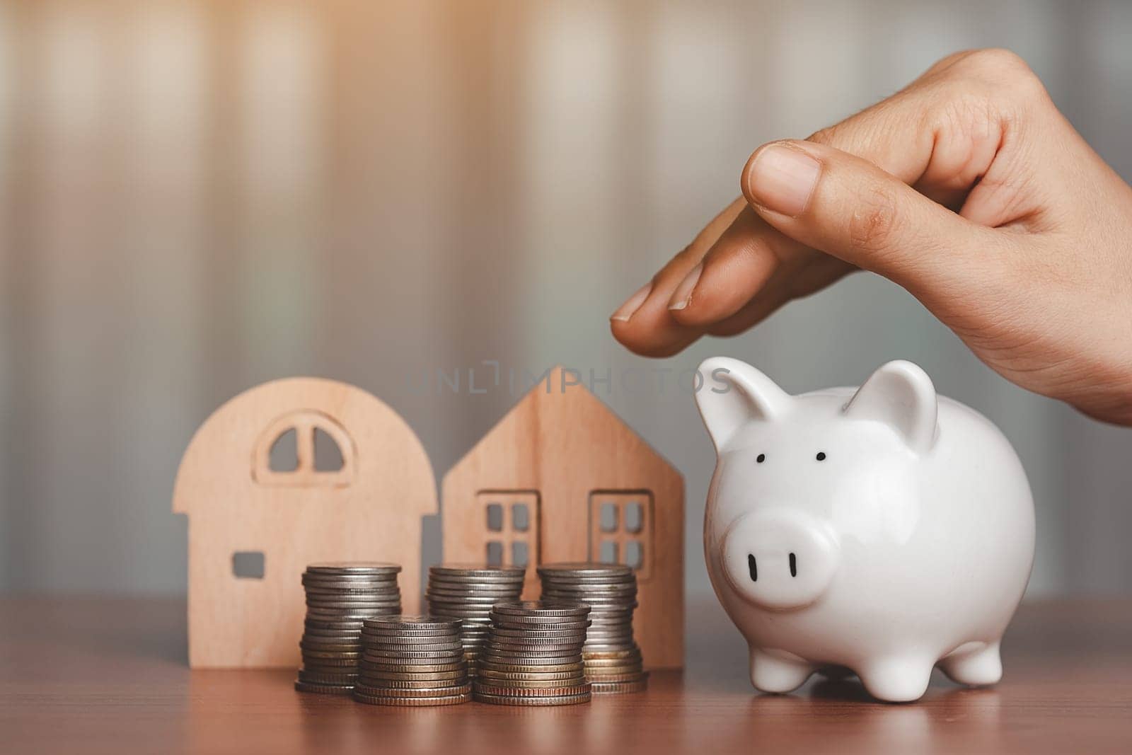 Hand covering a piggy bank with the wooden house model and a pile of coins and money on the table for business, finance, saving money and property investment concept.